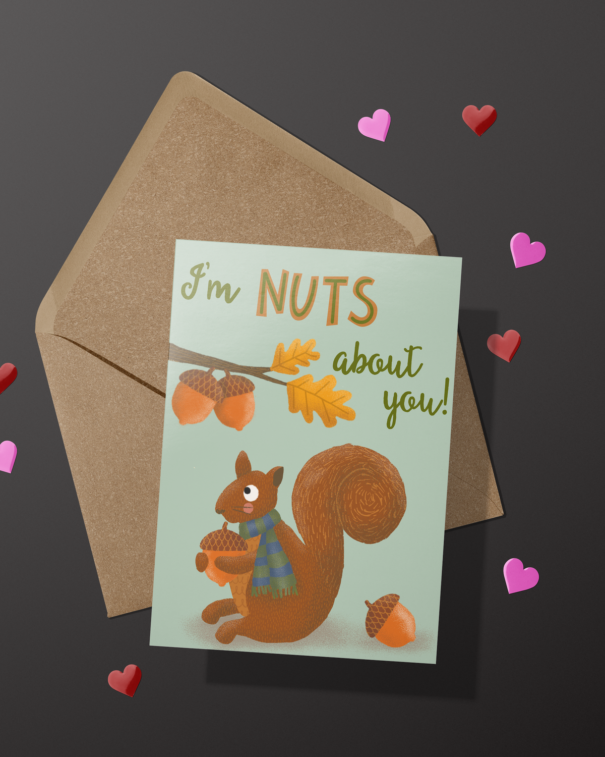 A greeting card with an illustration of a squirrel holding an acorn and the words I'm Nuts About You! on a kraft envelope on a black background with red and pink foil hearts