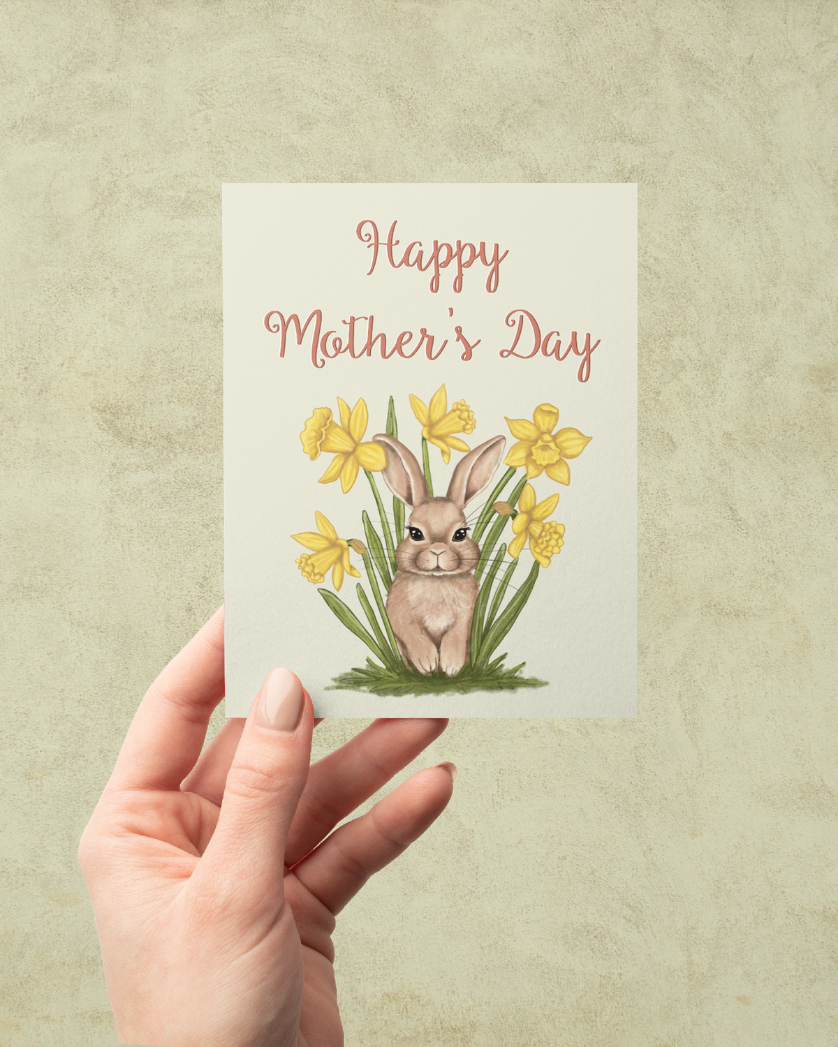 A hand holding a greeting card with a hand-painted bunny in daffodils and the words Happy Mother's Day against a muted background