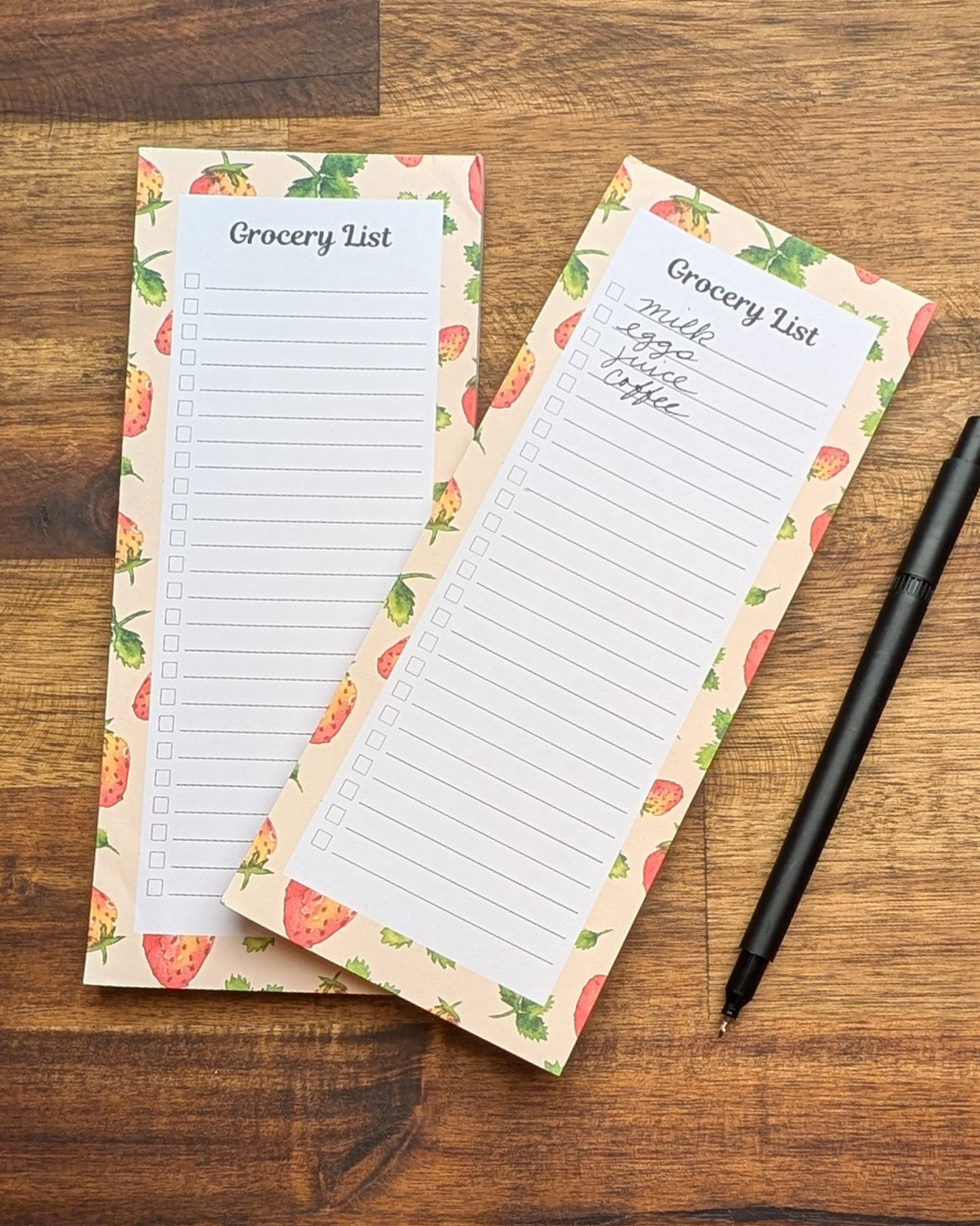 Two checklist grocery list notepads with a hand-painted watercolor strawberry border on a wood table with a black pen