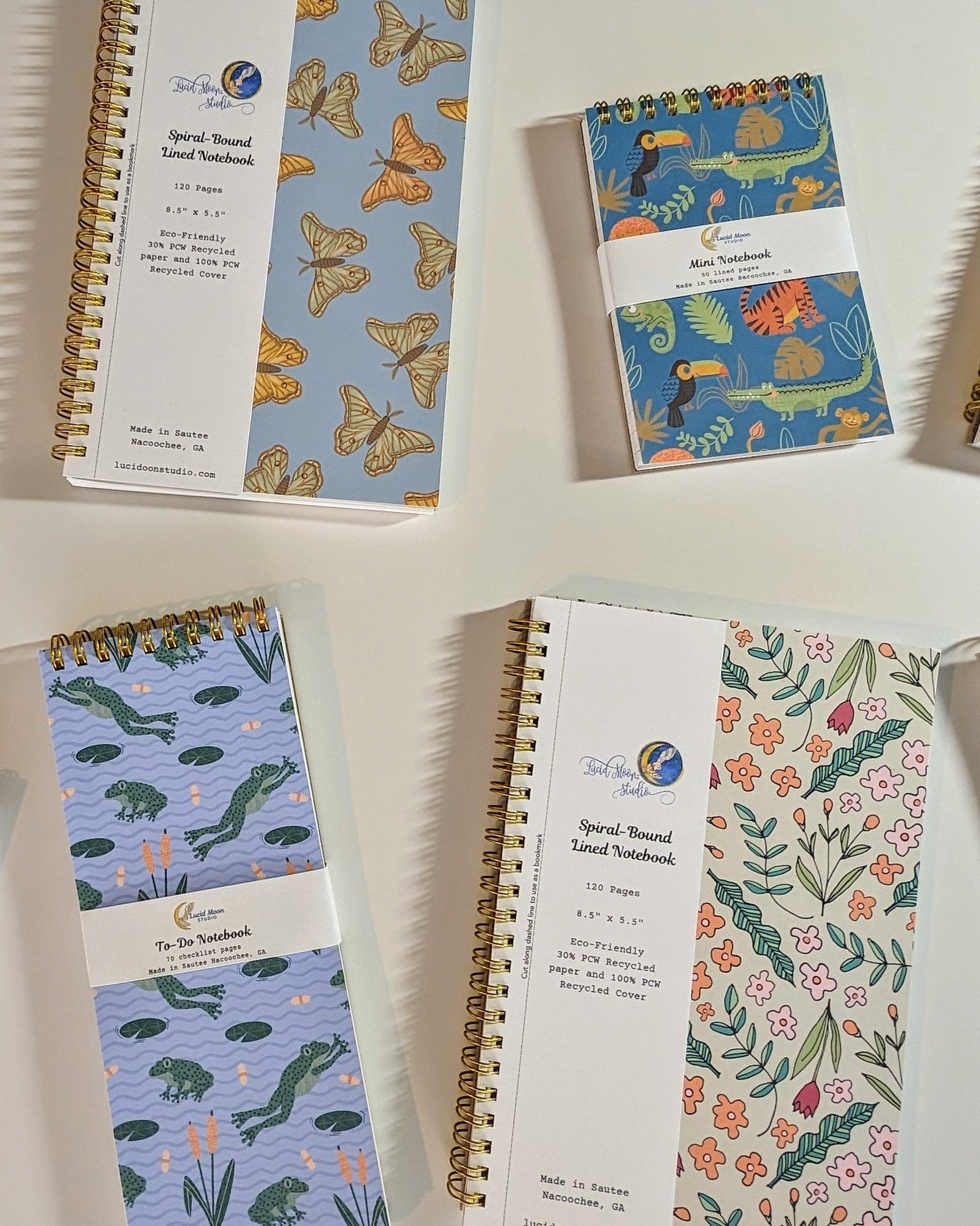 An assortment of Lucid Moon Studio spiral notebooks with colorful nature-inspired patterns on the covers on a white background