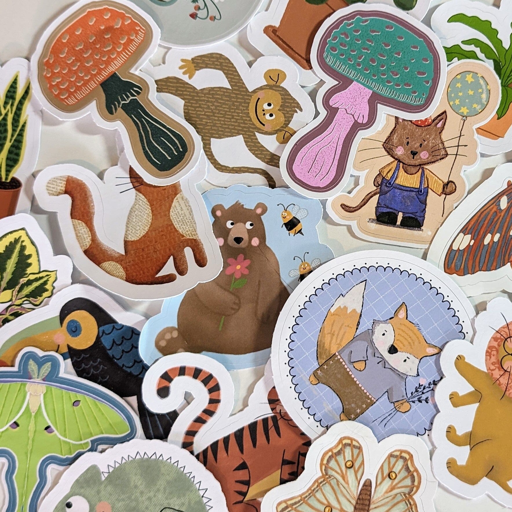 A photo of a huge assortment of cute die-cut glossy and matte waterproof vinyl stickers featuring mushrooms, cats, bears, foxes, monkeys, plants, butterflies, lions, toucans, iguanas, and tigers.