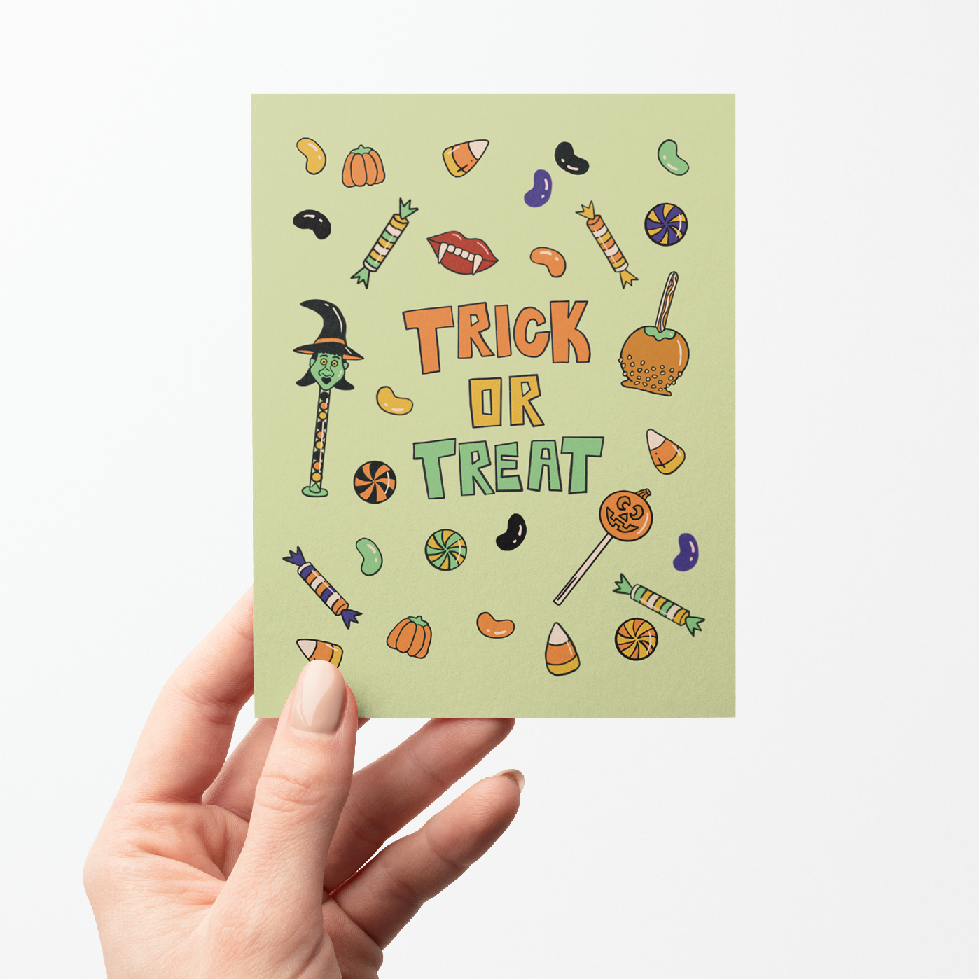 A hand holding a light sage green greeting card with a 'Trick or Treat' Halloween design, set against a plain white background.