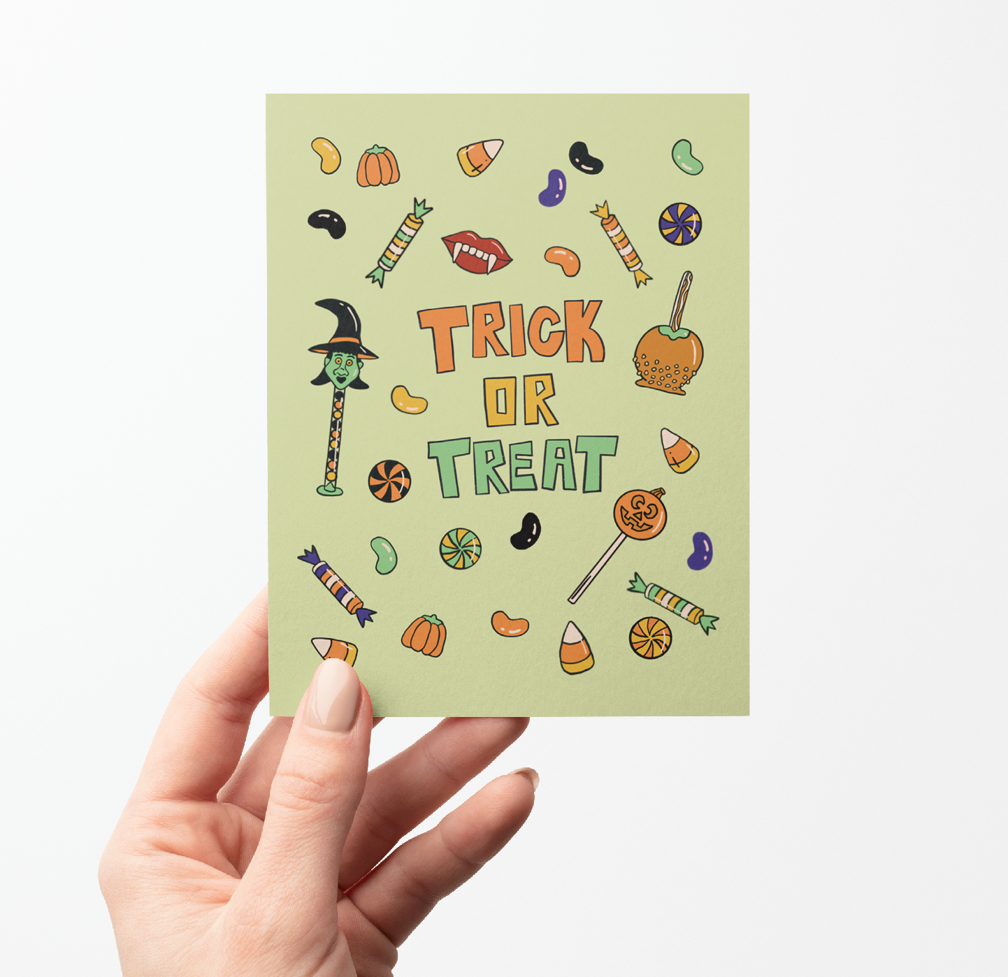 A hand holding a light sage green greeting card with a 'Trick or Treat' Halloween design, set against a plain white background.
