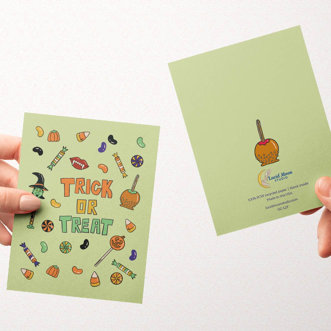 Two hands hold up a light green greeting card and its back side against a plain white background. The front of the card features colorful Halloween-themed illustrations and the words 'TRICK OR TREAT' in orange.