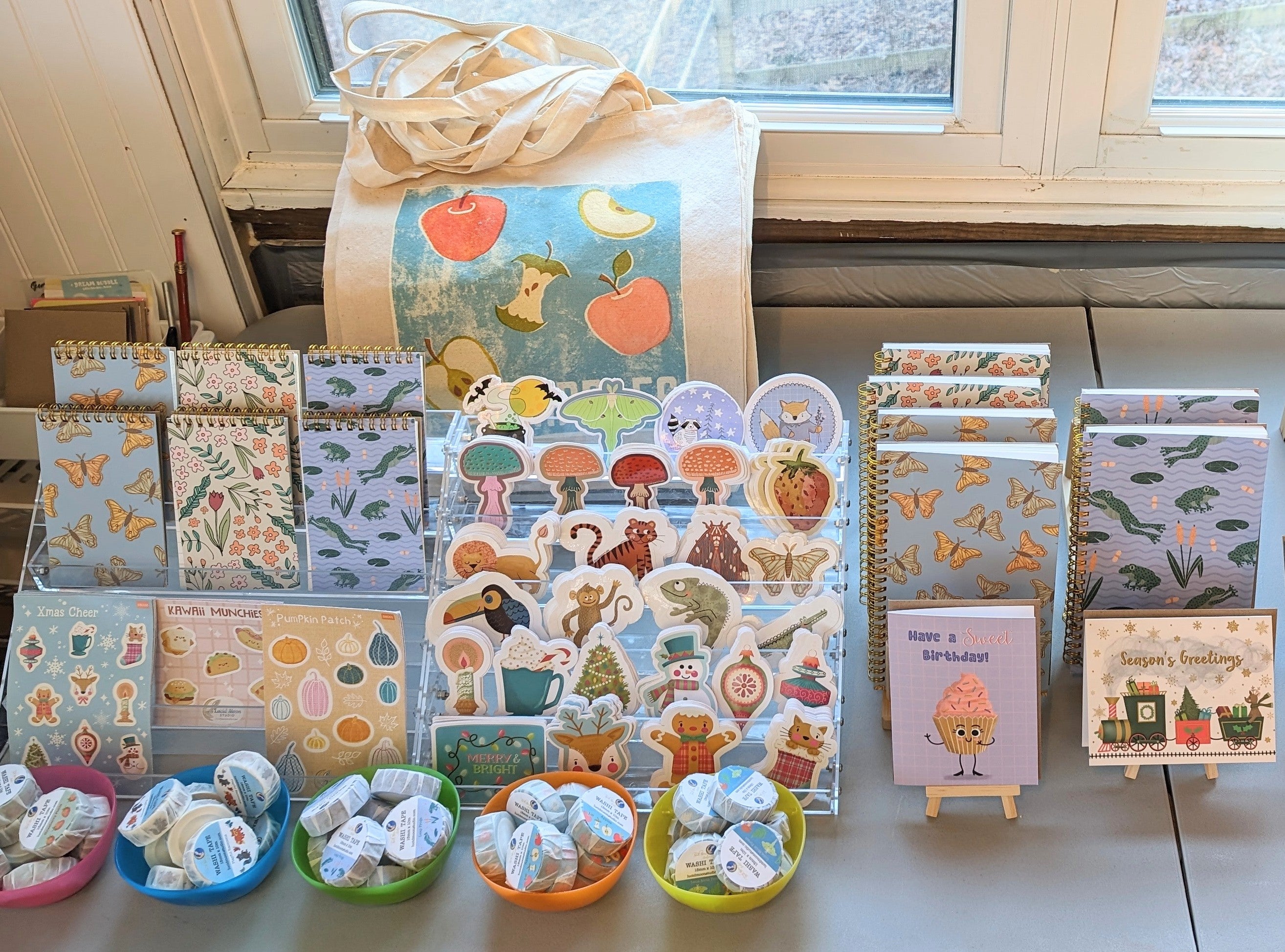 A photo of Lucid Moon Studio's sustainably made small batch products on a table that include sticker sheets, vinyl stickers, eco-friendly spiral-bound notebooks and jotters, washi tape, greeting cards, and tote bags all featuring designs by artist Lisa Petrillo..