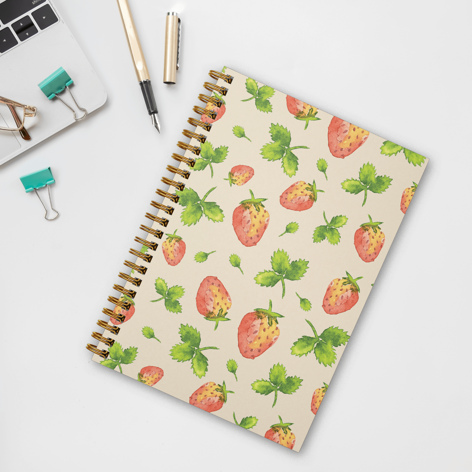 A photo of a wire spiral-bound notebook featuring watercolor-painted strawberries and leaves in a tossed pattern on the recycled cover laying on a white desk with clips, a pen, glasses, and part of a laptop showing.