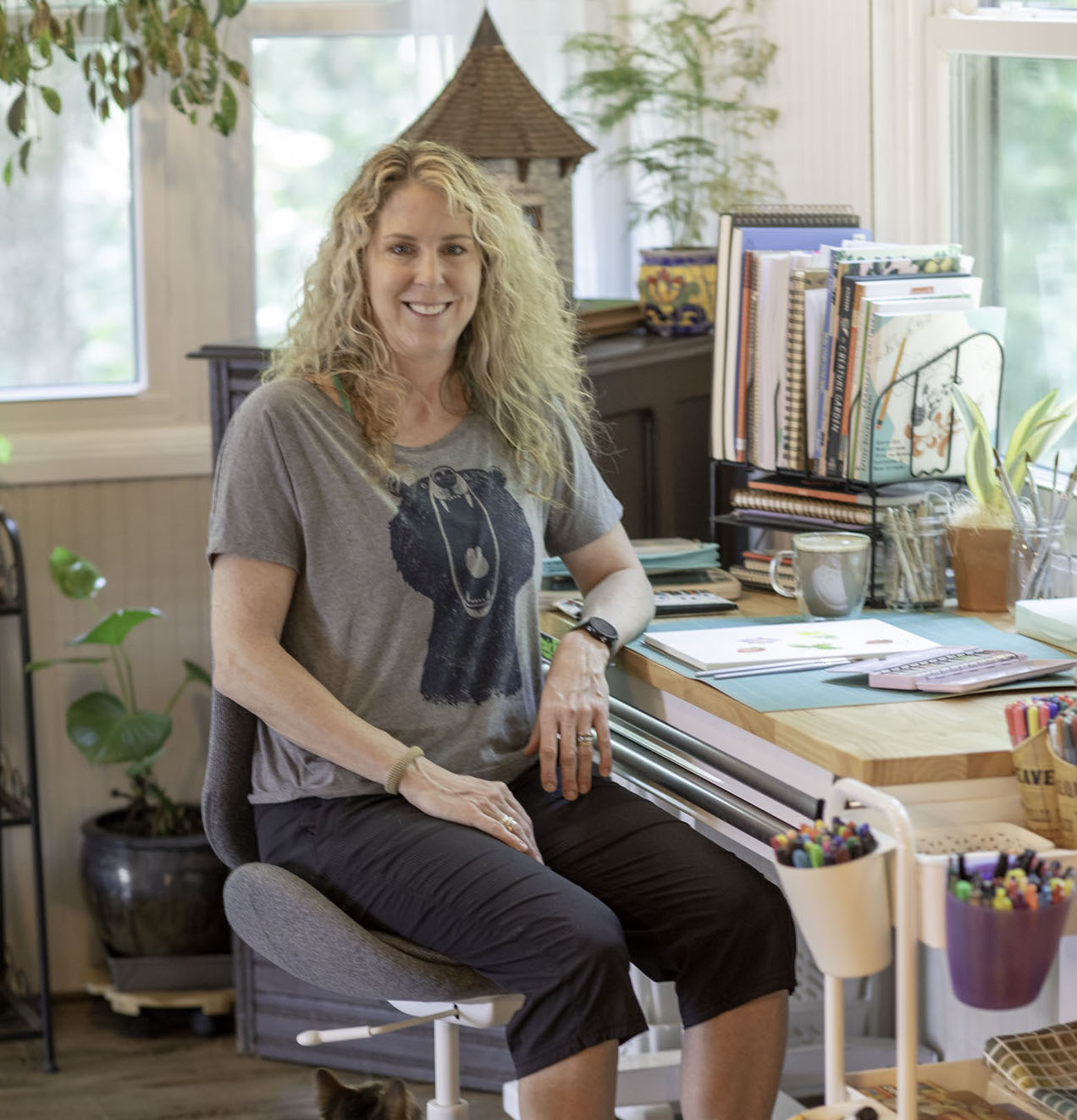 A photo of artist Lisa Petrillo in her studio in North Georgia sitting at a desk with art supplies, a painting, and plants in the background.