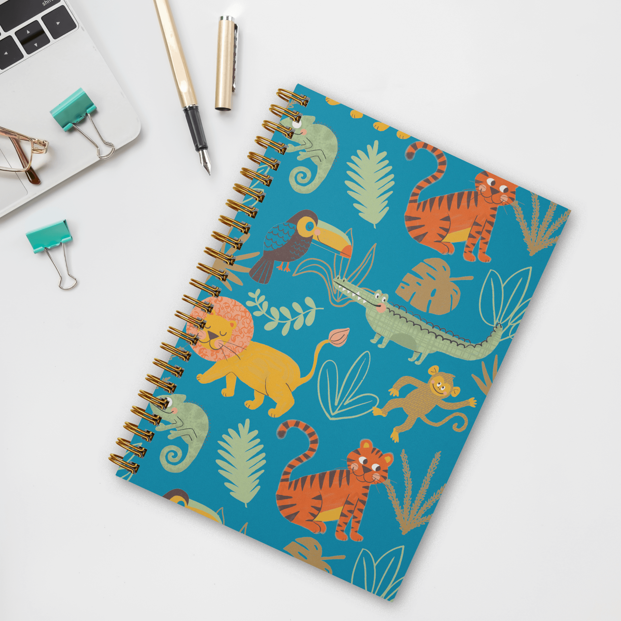 A photo of a wire spiral-bound notebook featuring hand-illustrated cute jungle animals such as lions, tigers, toucans, iguanas, monkeys, and crocodiles on a bright blue background with green and brown leaves in a tossed pattern on the recycled cover layin