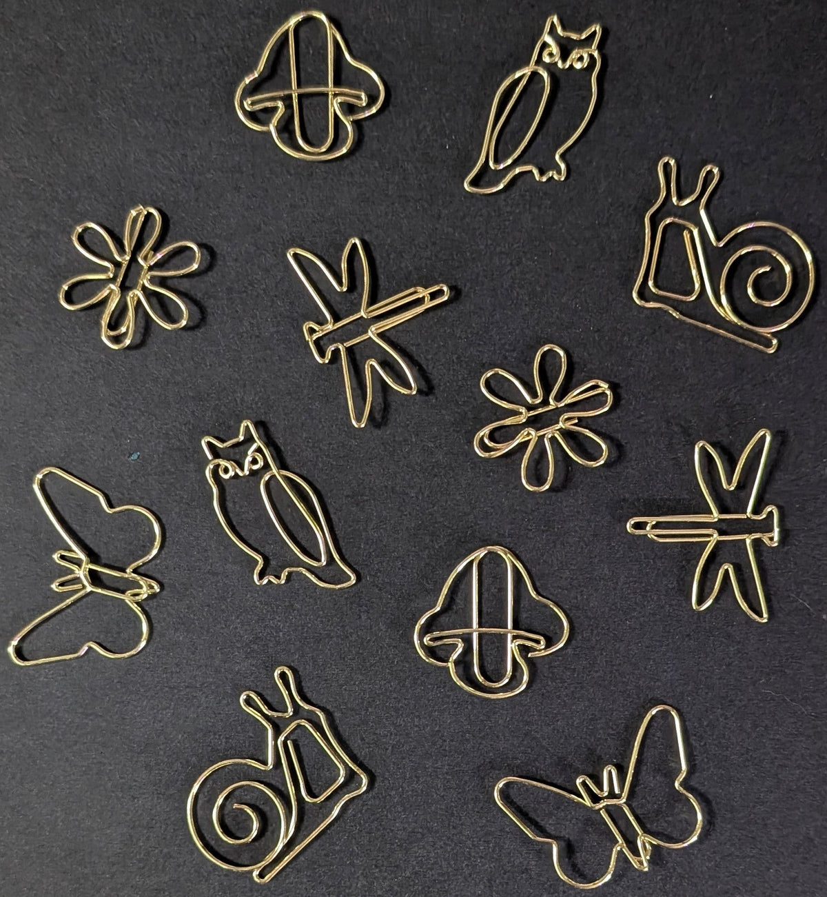 A set of small gold metal wire paper clips shaped like owls, butterflies, snails, flowers, and a clover, arranged on a dark textured background.