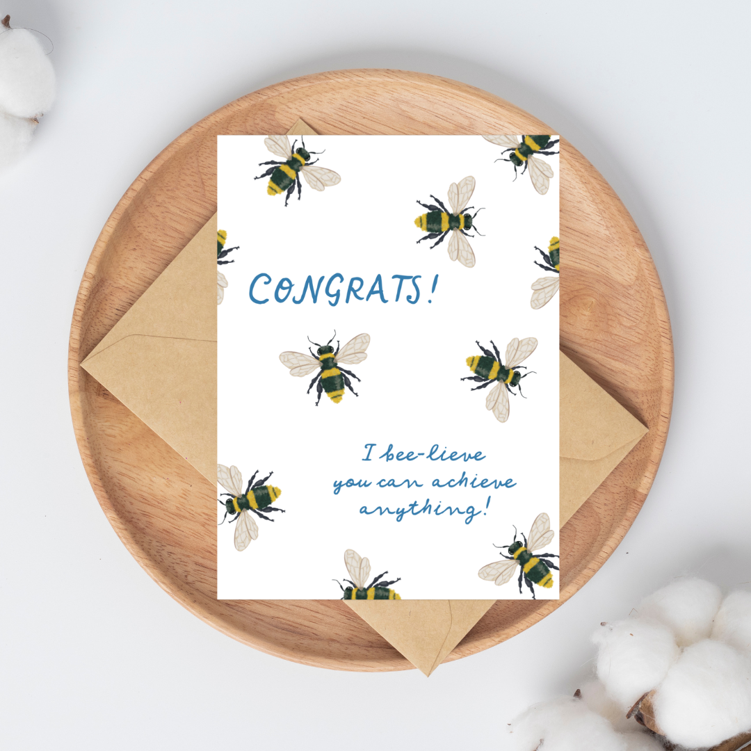 Greeting card with hand-drawn bee illustrations on a white background with blue text that says "Congrats! I bee-lieve you can achieve anything!" with a brown kraft envelope.