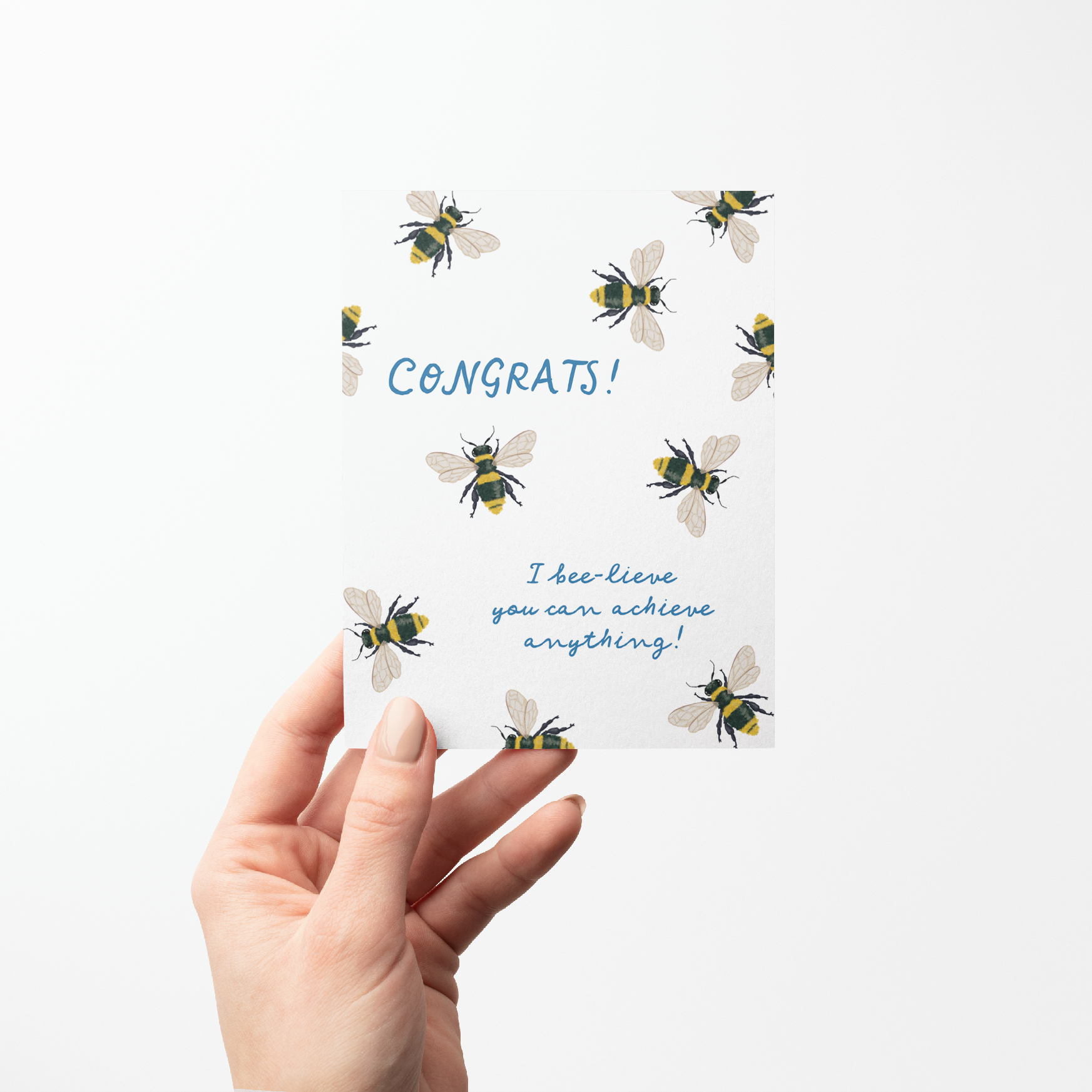 Hand holding a Greeting card with hand-drawn bee illustrations on a white background with blue text that says "Congrats! I bee-lieve you can achieve anything!"