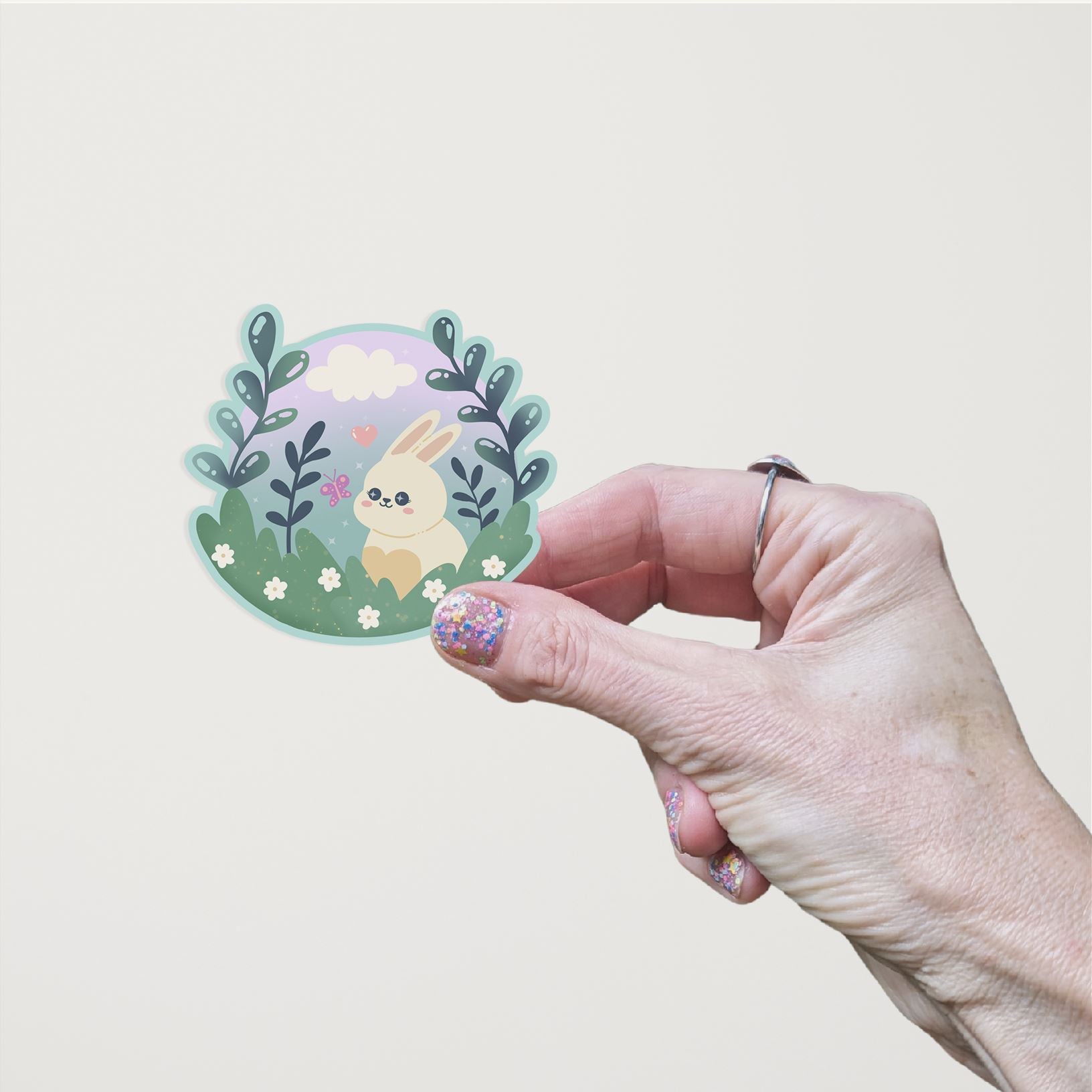 A hand holding a hand-drawn die cut matte vinyl sticker featuring a cute Kawaii bunny sitting in a meadow surrounded by leaves, flowers, a pink butterfly, a heart, and a cloud in the background.