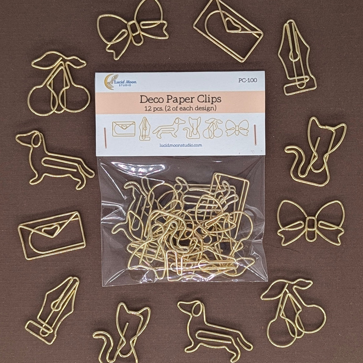 A set of 12 gold-colored metal wire paper clips shaped like cherries, bows, envelopes, a fountain pen, a dog, a squirrel, and a cherry, arranged around a clear plastic bag of the product on a solid brown background.