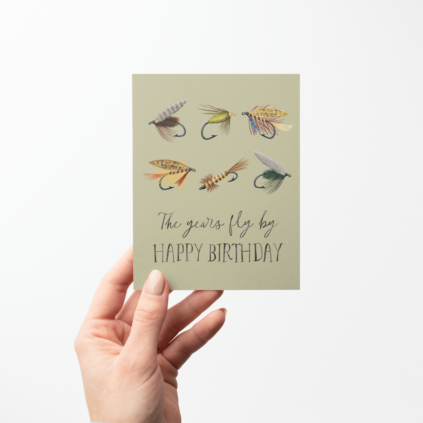 Hand holding a Birthday card with 6 fishing fly illustrations and text "The years fly by HAPPY BIRTHDAY" on a light green background.