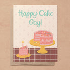 A photo of a recycled eco-friendly A2 greeting card with a hand-drawn illustration of a pink layered cake on a cake stand and a slice of cake with a candle in it on a plate on a tiled countertop with the phrase," Happy Cake Day!" on the front on top of a