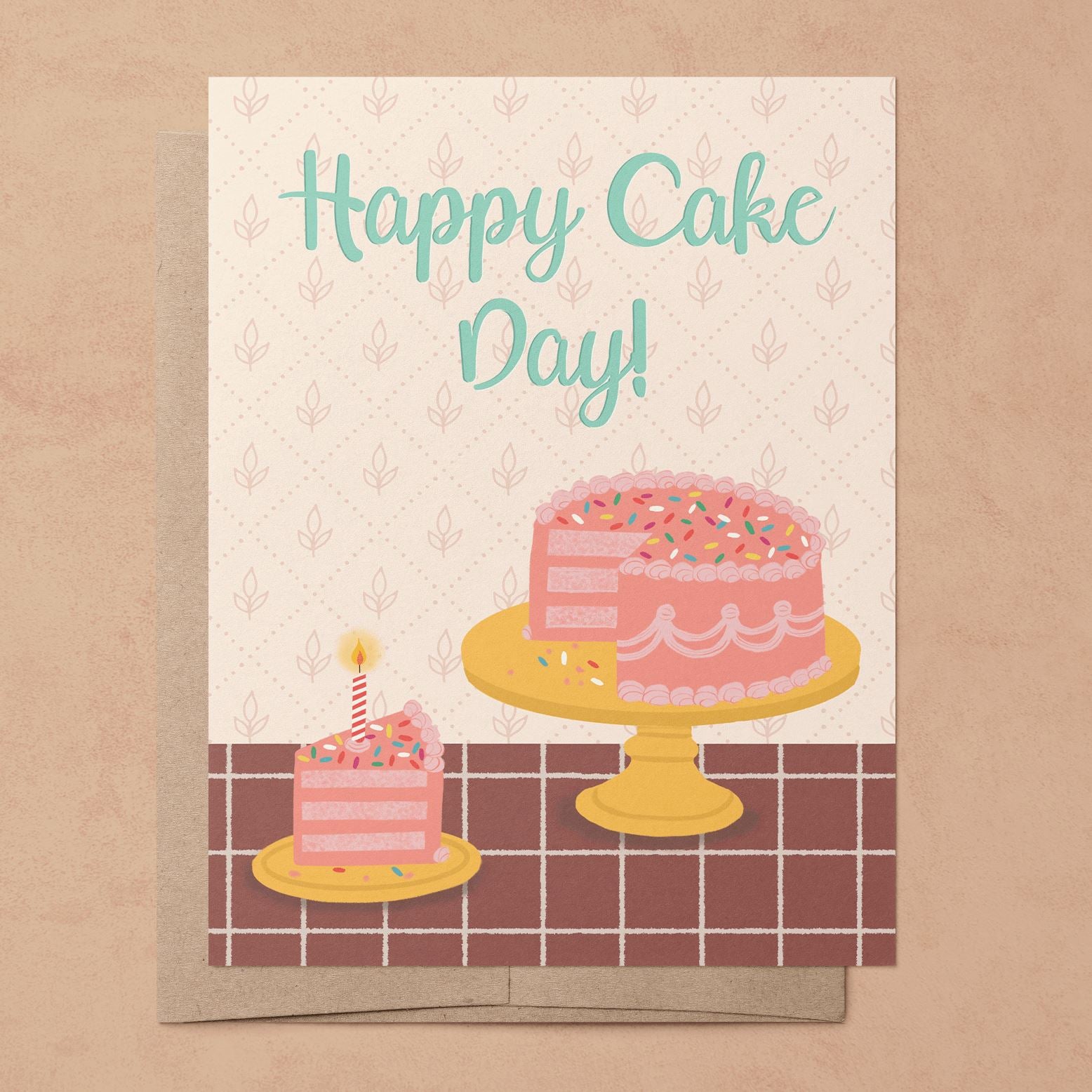 A photo of a recycled eco-friendly A2 greeting card with a hand-drawn illustration of a pink layered cake on a cake stand and a slice of cake with a candle in it on a plate on a tiled countertop with the phrase," Happy Cake Day!" on the front on top of a 