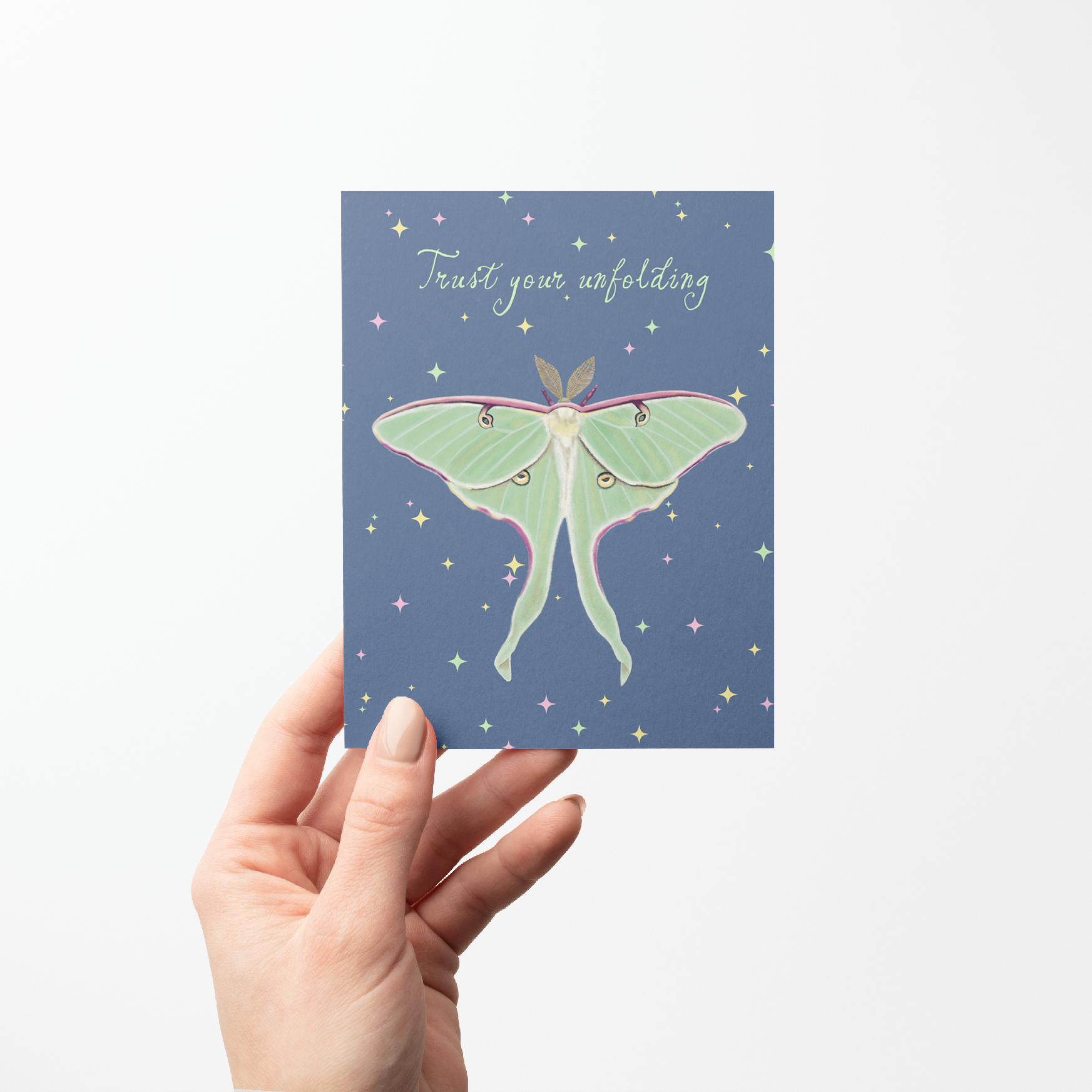 Hand holding a Greeting card with a Luna moth illustration and "Trust your unfolding" text on a blue background with colored stars on it.