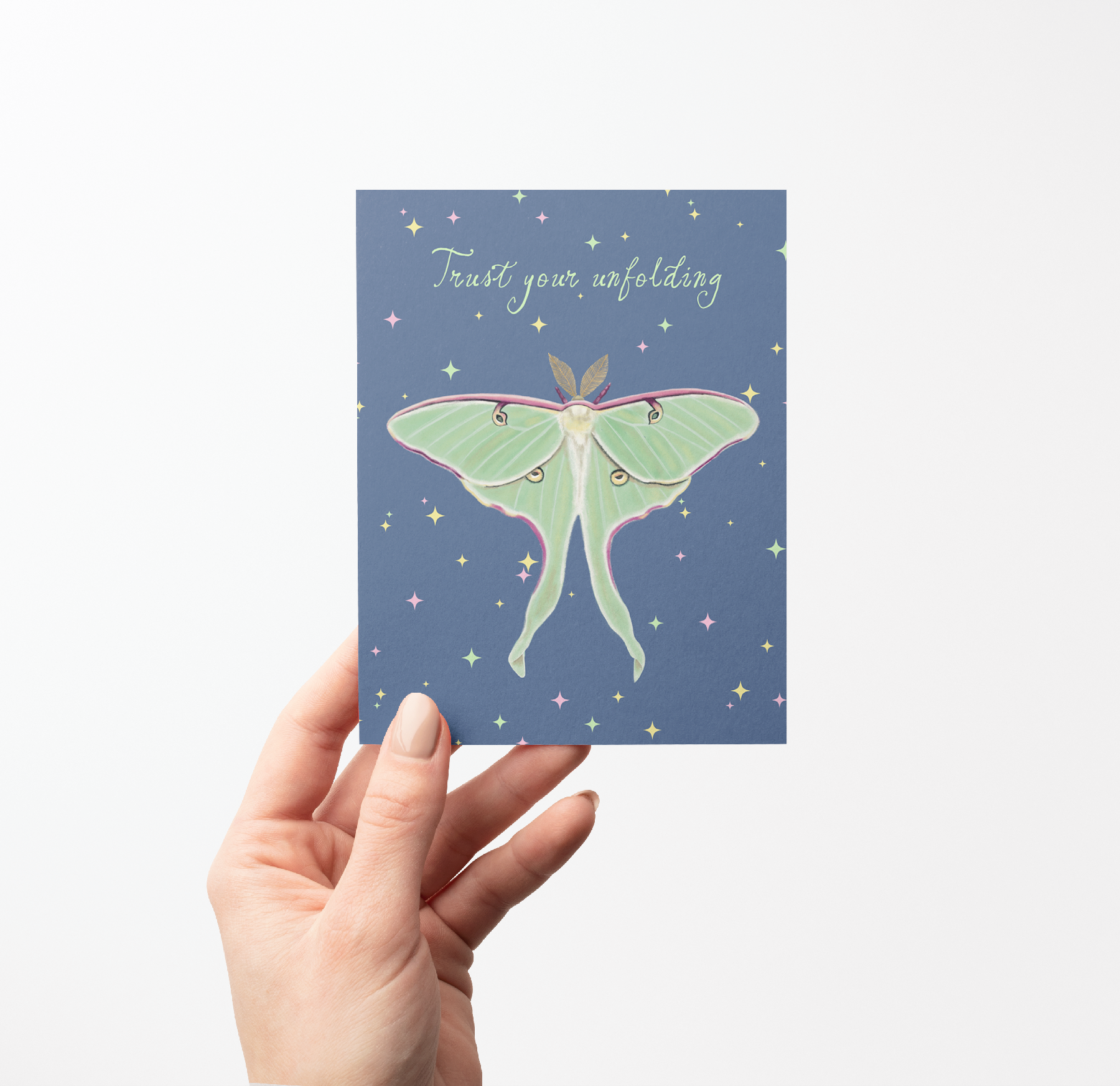 Hand holding a Greeting card with a Luna moth illustration and "Trust your unfolding" text on a blue background with colored stars on it.