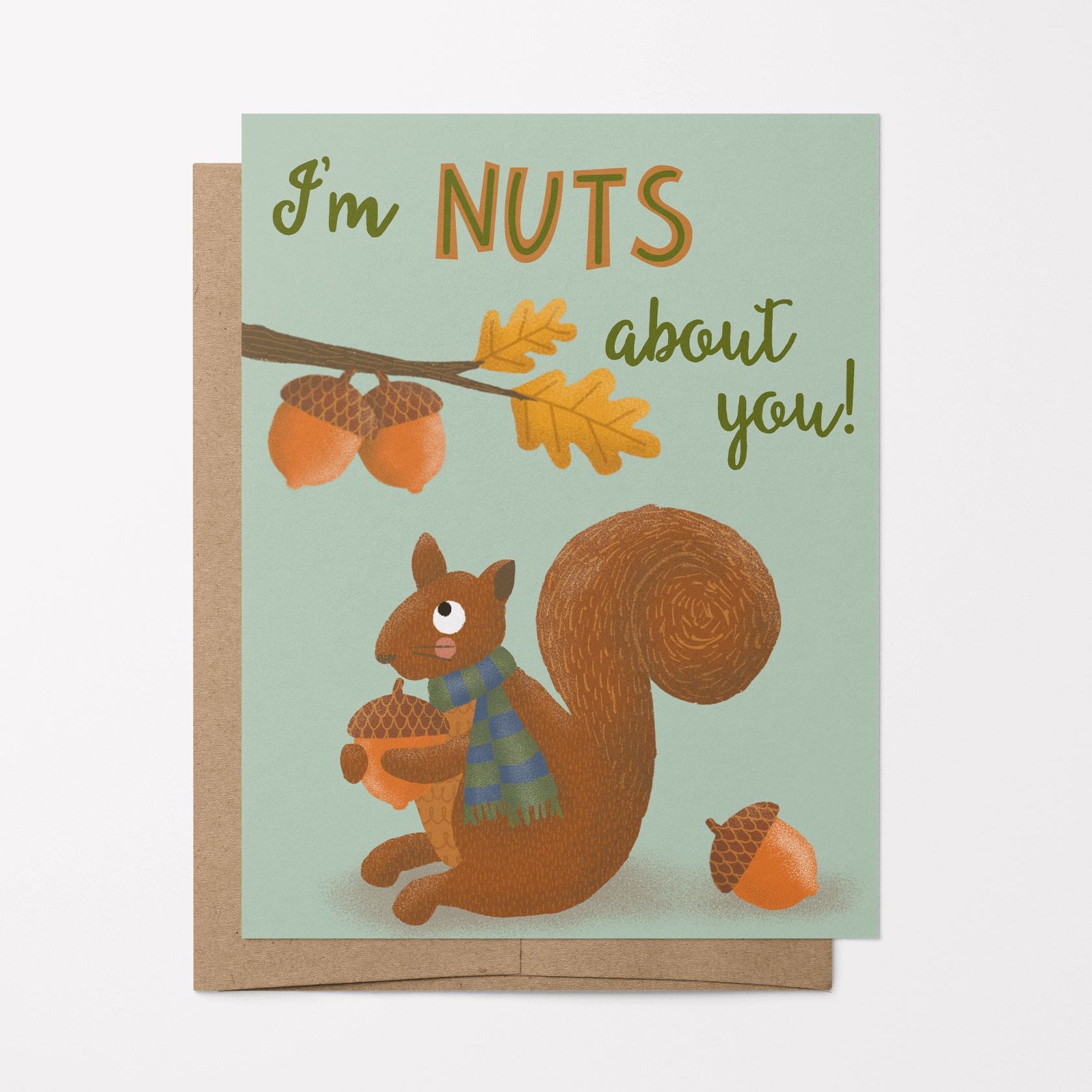 A photo of greeting card and brown kraft envelope featuring a hand-drawn illustration of a cute squirrel with a scarf on holding an acorn looking at a branch with acorns and the phrase, "I'm nuts about you!"