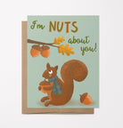 A photo of greeting card and brown kraft envelope featuring a hand-drawn illustration of a cute squirrel with a scarf on holding an acorn looking at a branch with acorns and the phrase, "I'm nuts about you!"
