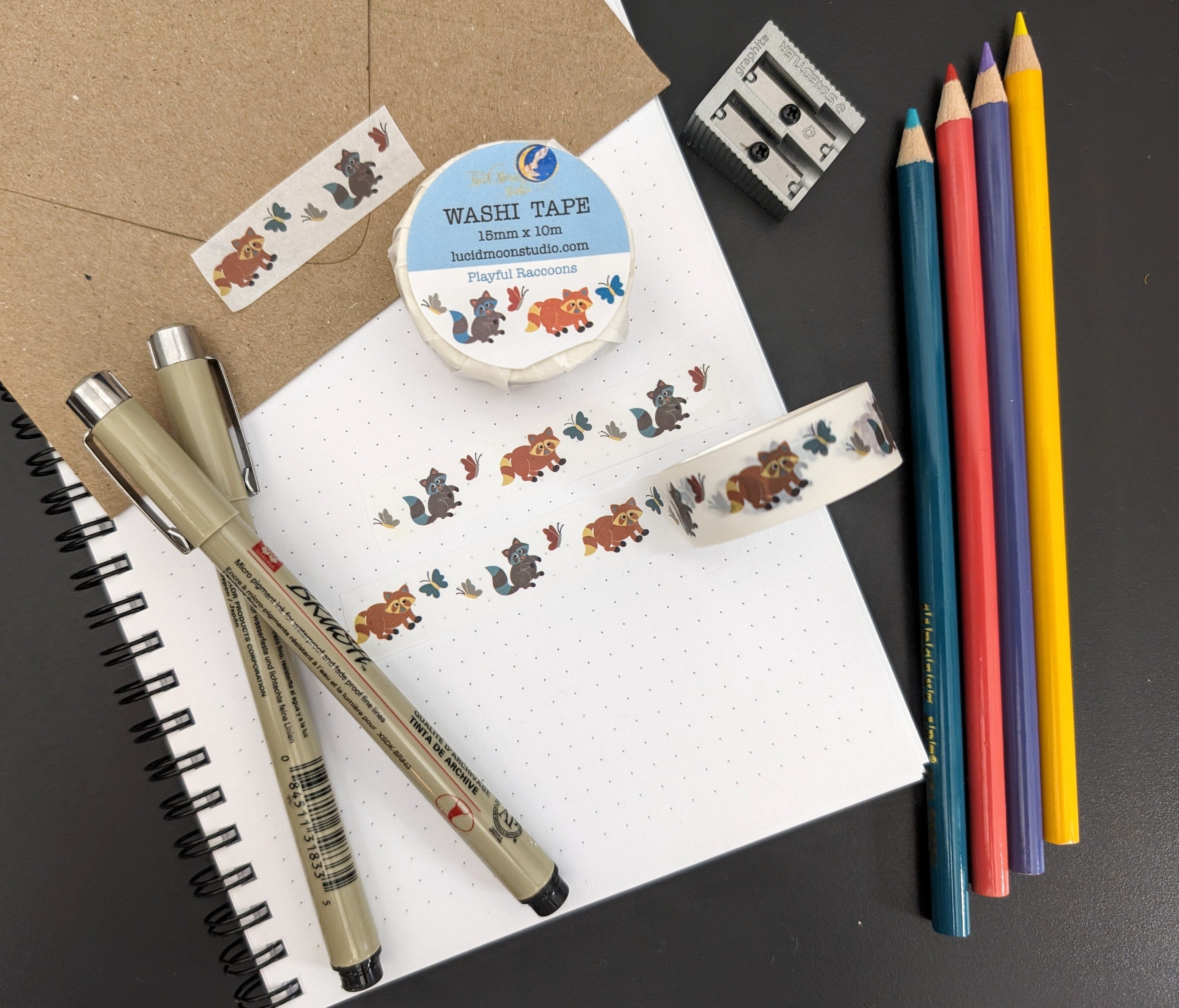 A roll of washi tape in a paper wrapper and 2 strips of washi tape adorned with hand drawn brown and gray raccoons and colorful butterflies on a white background on a notebook and kraft envelope along with pens, colored pencils, and a pencil sharpener.