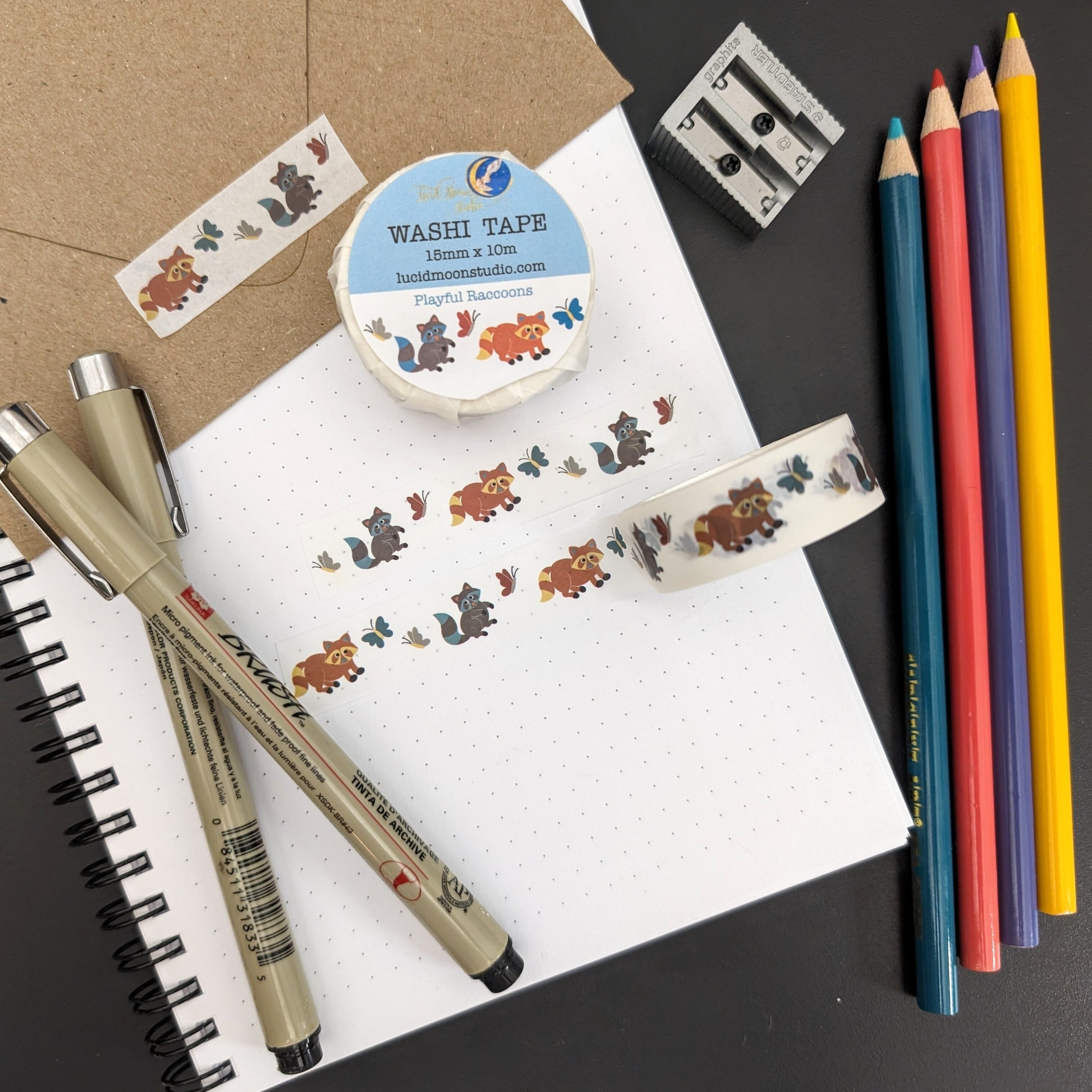 A roll of washi tape in a paper wrapper and 2 strips of washi tape adorned with hand drawn brown and gray raccoons and colorful butterflies on a white background on a notebook and kraft envelope along with pens, colored pencils, and a pencil sharpener.