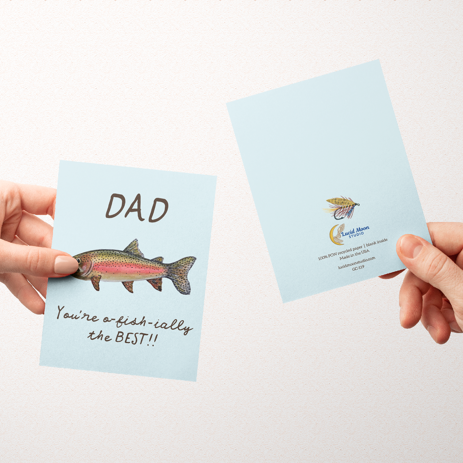 Two hands holding greeting cards with the front and back of a Greeting card with a watercolor rainbow trout illustration and the text "Dad You're o-fish-ially the BEST" on a light blue background.