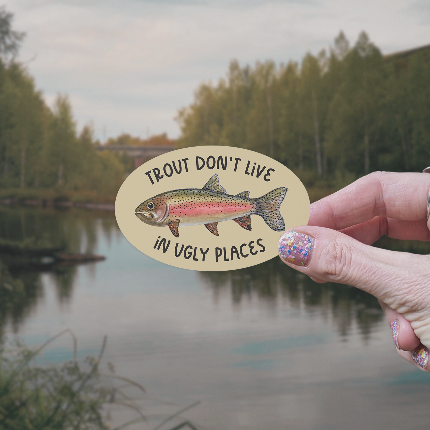 Hand holding an oval-shaped sticker with a watercolor rainbow trout illustration and the words "trout don't live in ugly places" on a tan background with a lake in the background.