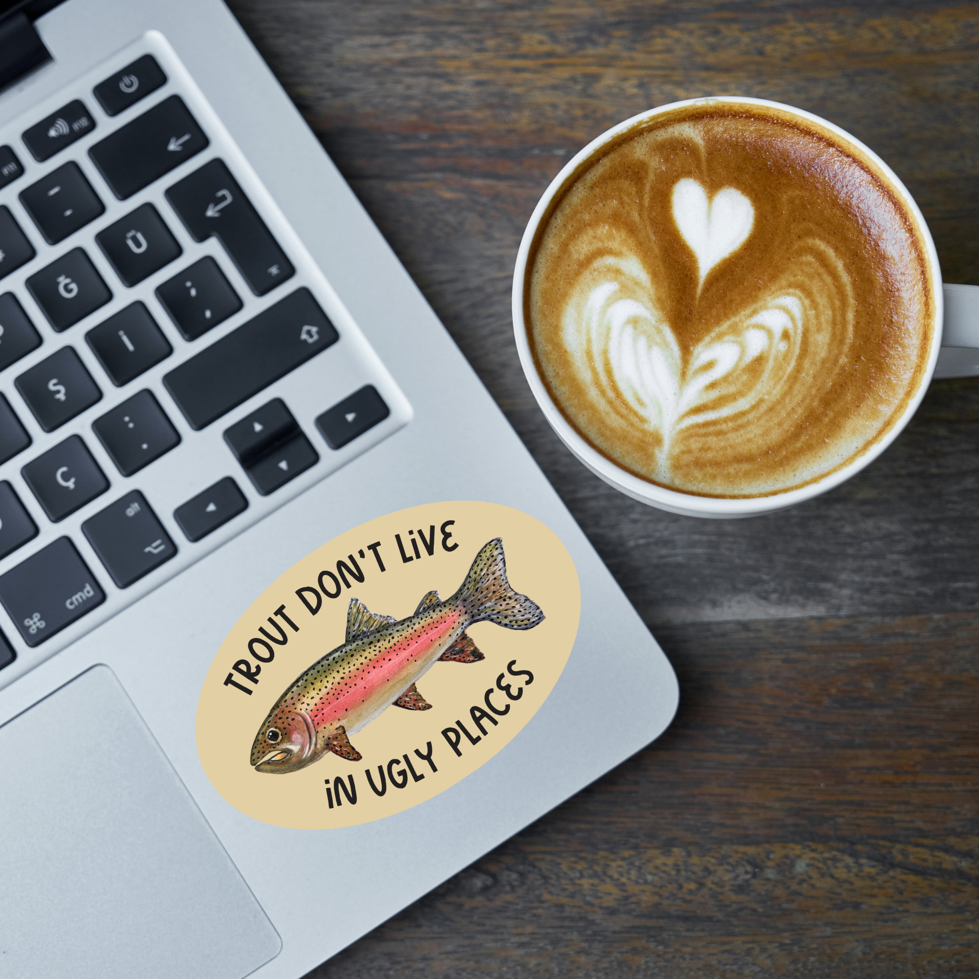 Laptop with an oval-shaped sticker with a watercolor rainbow trout illustration and the words "trout don't live in ugly places" on a tan background stuck on it next to a cup of coffee with latte art on a wooden surface.