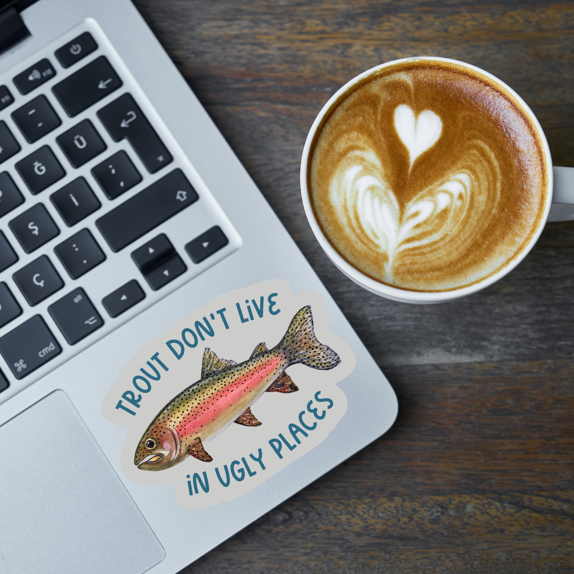 Laptop with a die cut sticker with a watercolor rainbow trout illustration and the text "Trout don't live in ugly places" on a grey background stuck on it next to a cup of coffee on a wooden surface