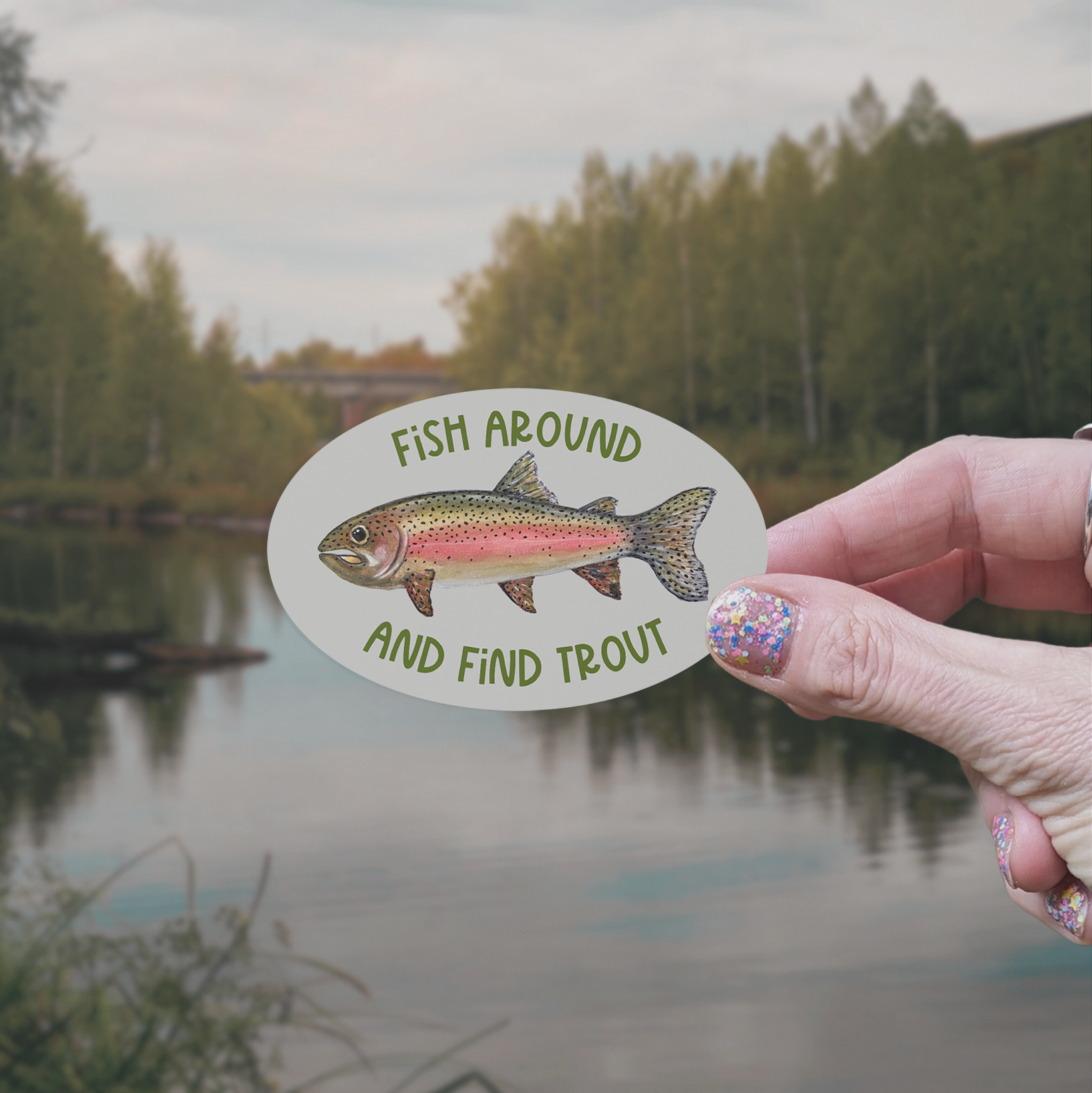 Hand holding an oval-shaped sticker with a watercolor rainbow trout illustration and green text "Fish Around and Find Trout" on a grey background with a lake in the background
