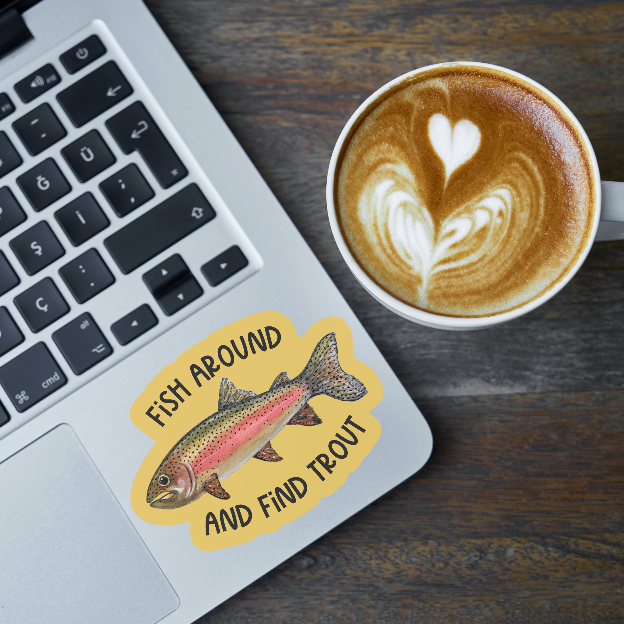 Laptop with a Die cut vinyl sticker with a watercolor rainbow trout illustration on a mustard-colored background with the words "Fish around and find trout" next to a cup of coffee on a wooden surface