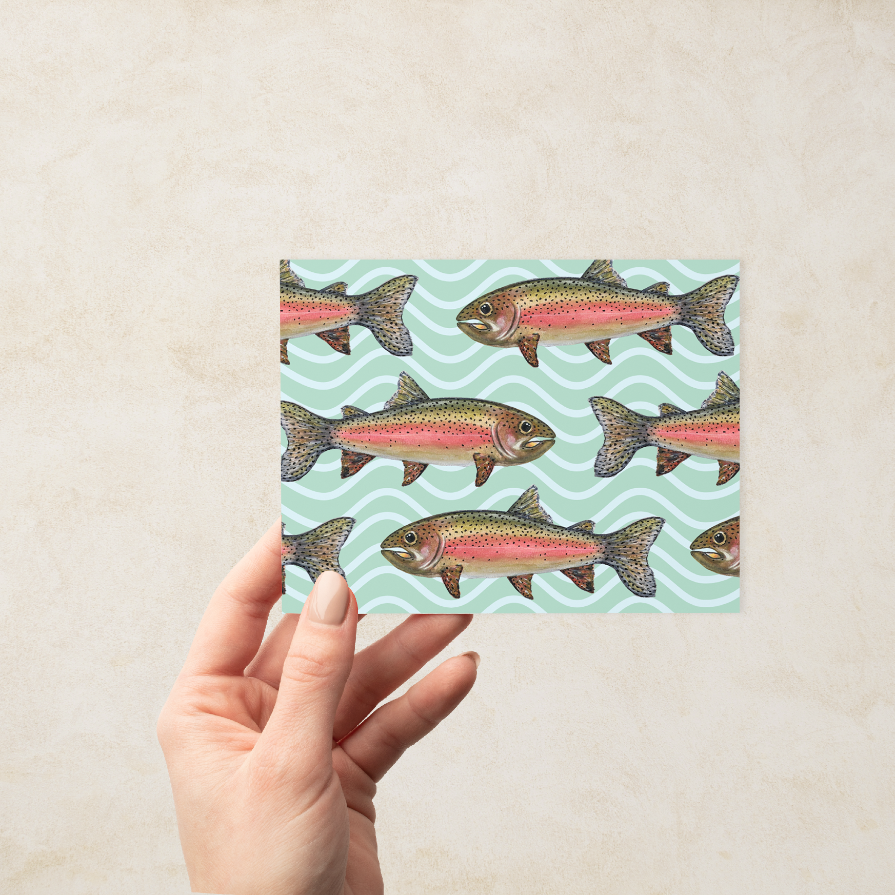 Hand holding a Greeting card with a school of watercolor rainbow trout illustrations on a mint green wavy pattern background.