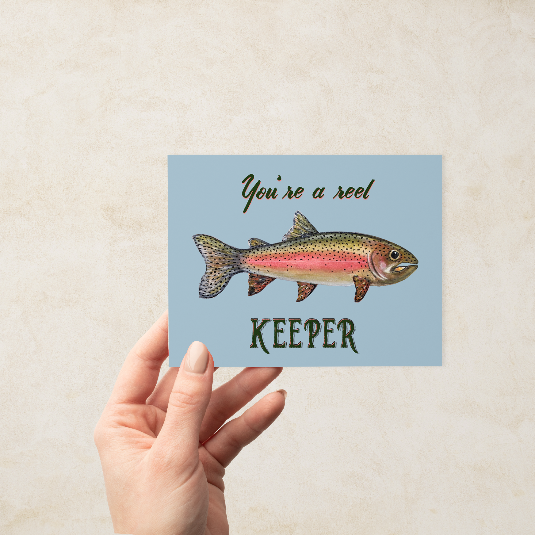 Hand holding a Greeting card with a watercolor rainbow trout illustration and text "You're a reel keeper" on a light blue background.