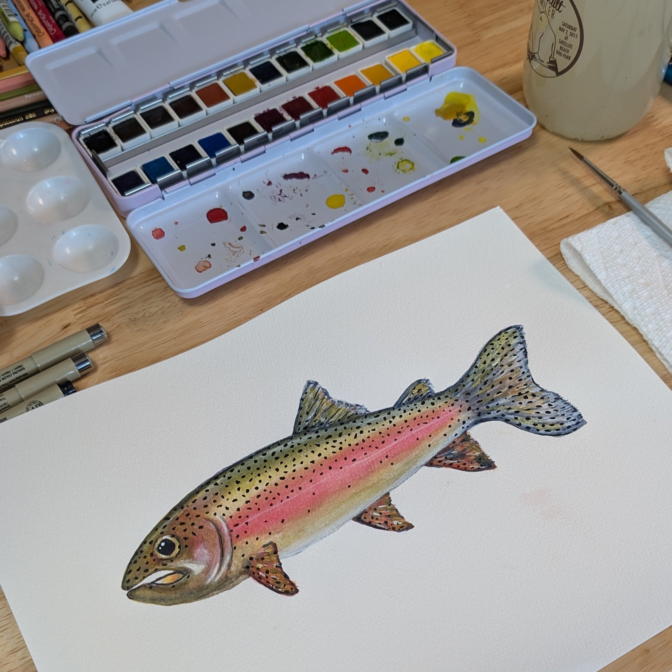 Watercolor painting of a rainbow trout on paper with watercolor set and brushes in the background.