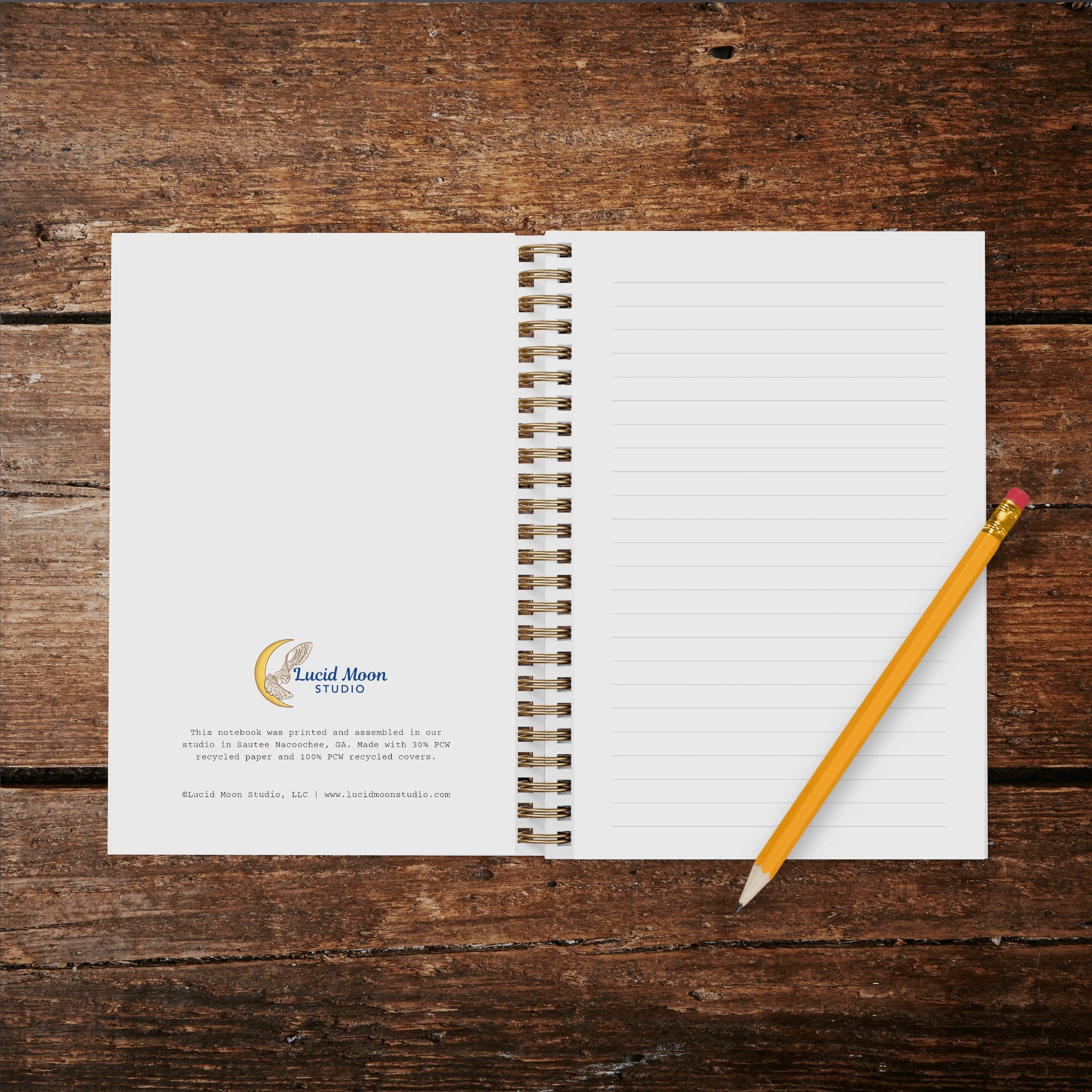 A photo of an opened wire spiral-bound notebook showing inside cover with Lucid Moon Studio logo and info about the notebook and lined pages on a wood background with a pencil.