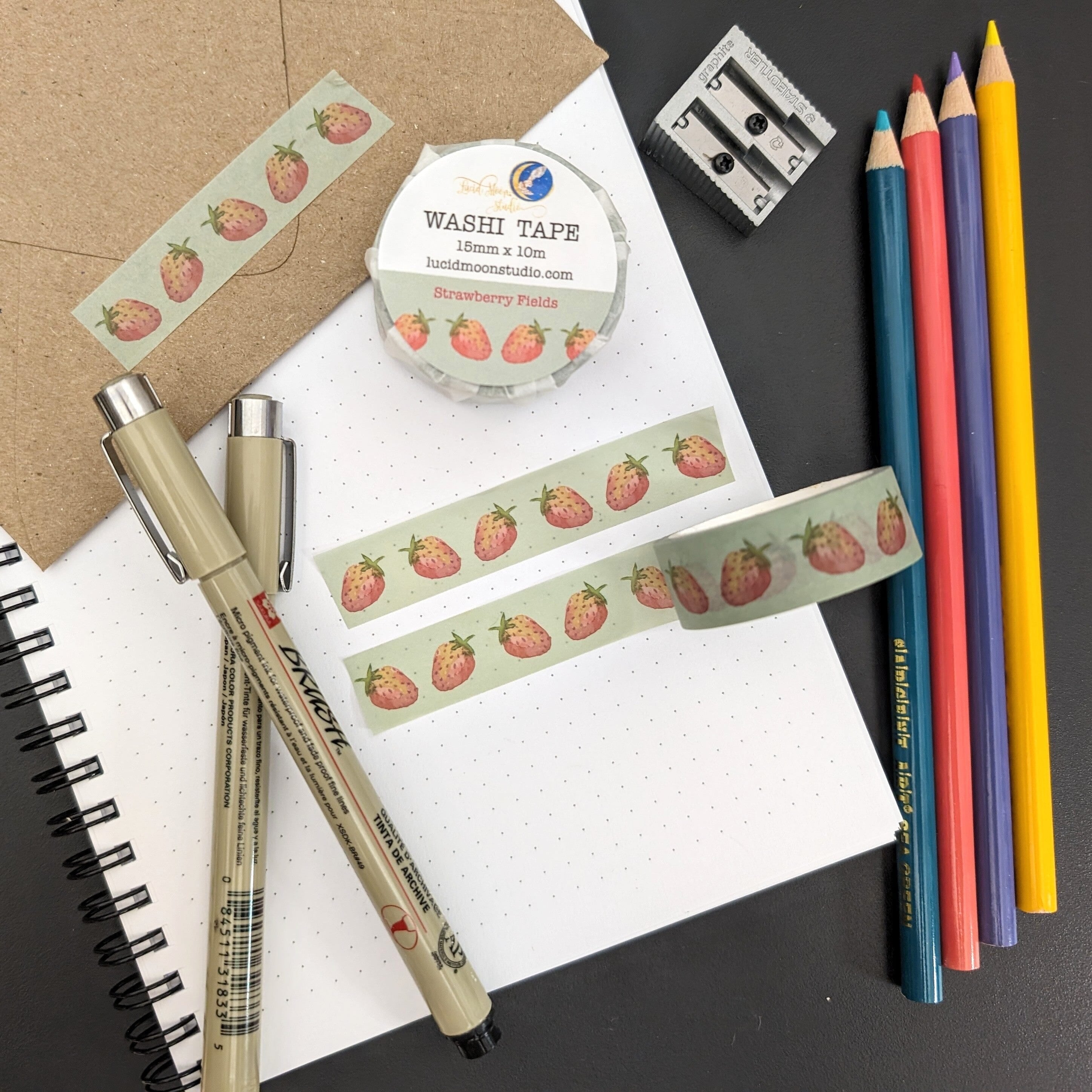 A roll of Strawberry Fields washi tape in a paper wrapper and 2 strips of the washi tape on a dot grid notebook and kraft envelope along with pens, colored pencils, and a pencil sharpener.