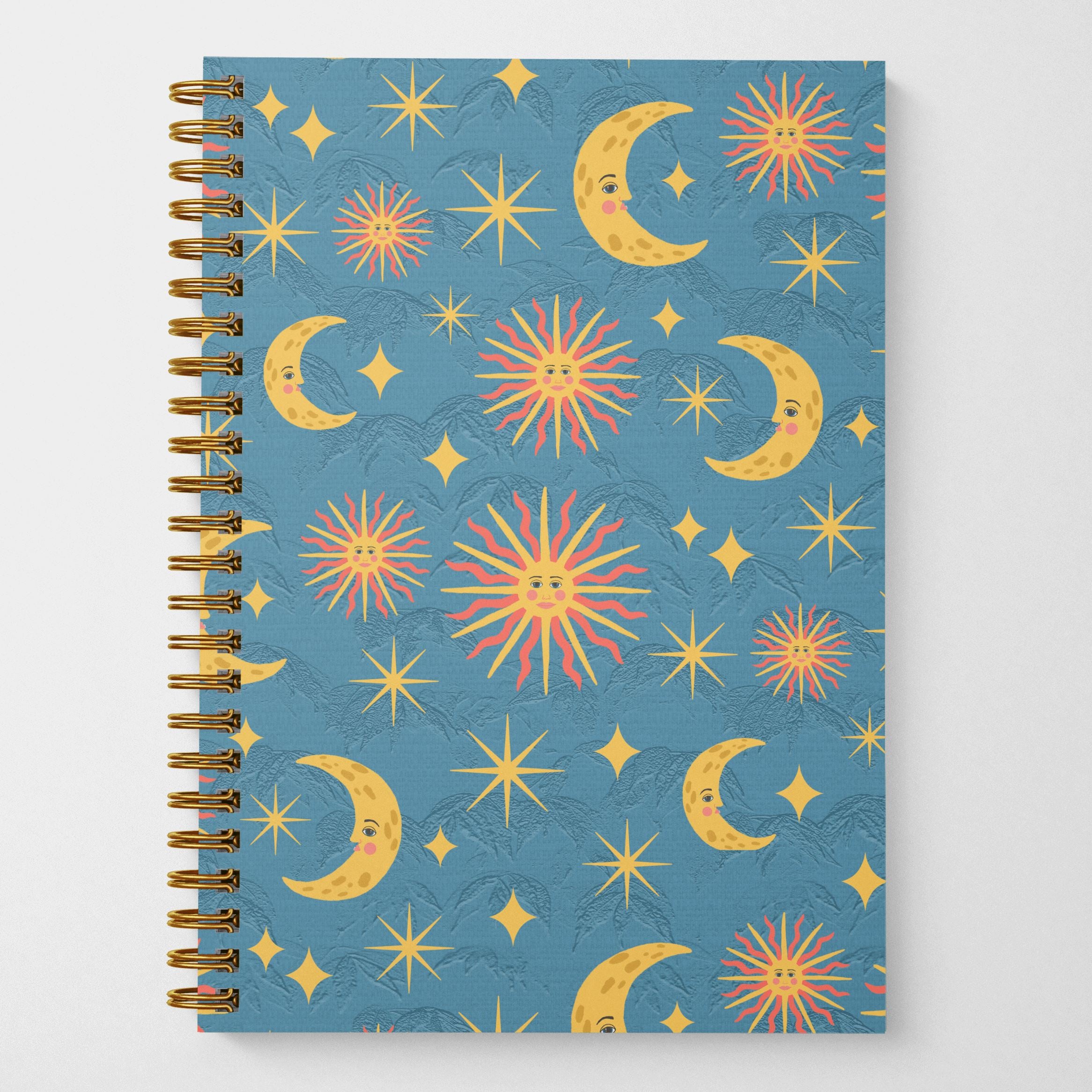 An eco friendly 8.5 x 5.5 inch wire spiral bound notebook with 120 lined pages featuring hand illustrated suns, moons, and stars pattern on a blue background on the cover.