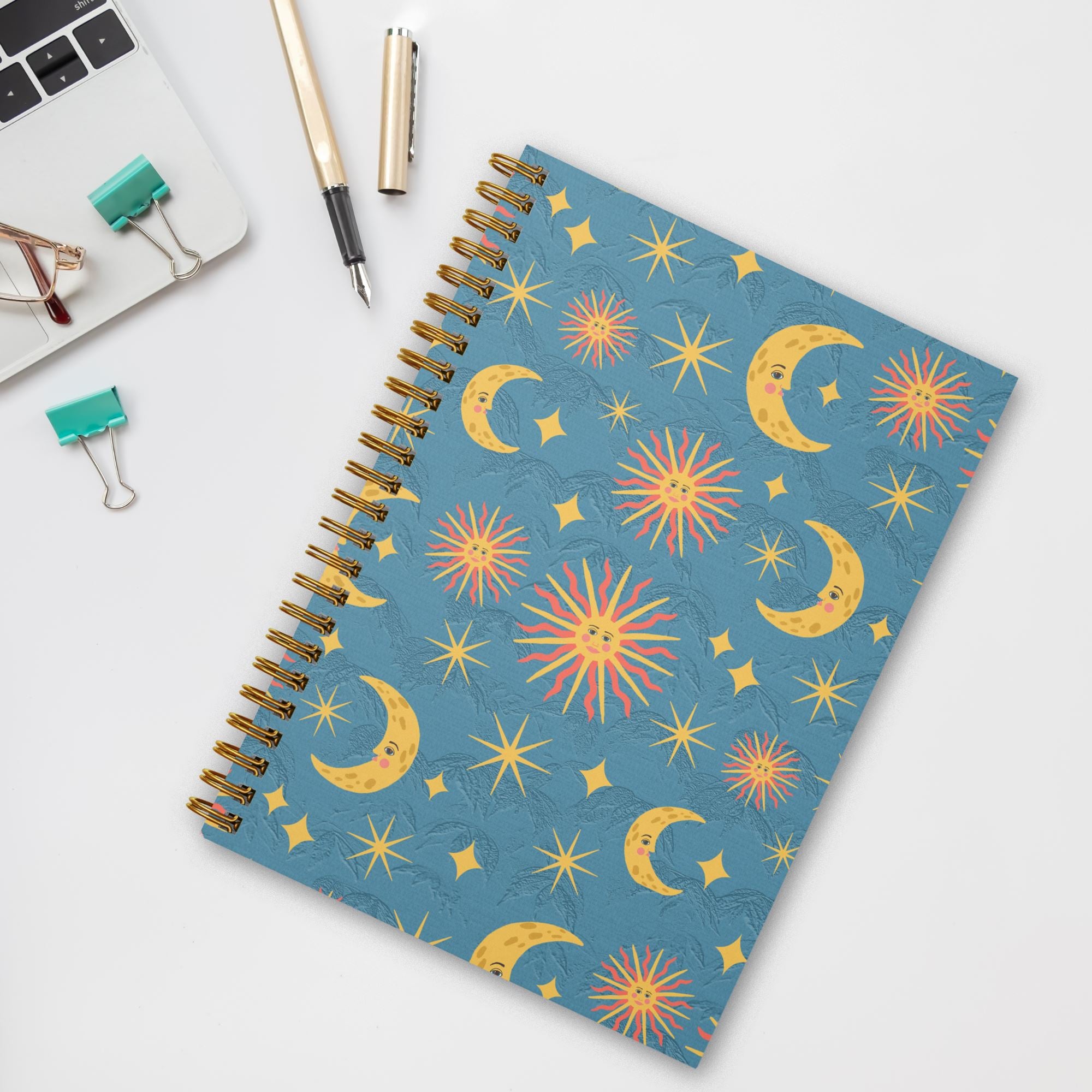 An eco friendly 8.5 x 5.5 inch wire spiral bound notebook with 120 lined pages featuring hand illustrated suns, moons, and stars pattern on a blue background on the cover on a table with office supplies.