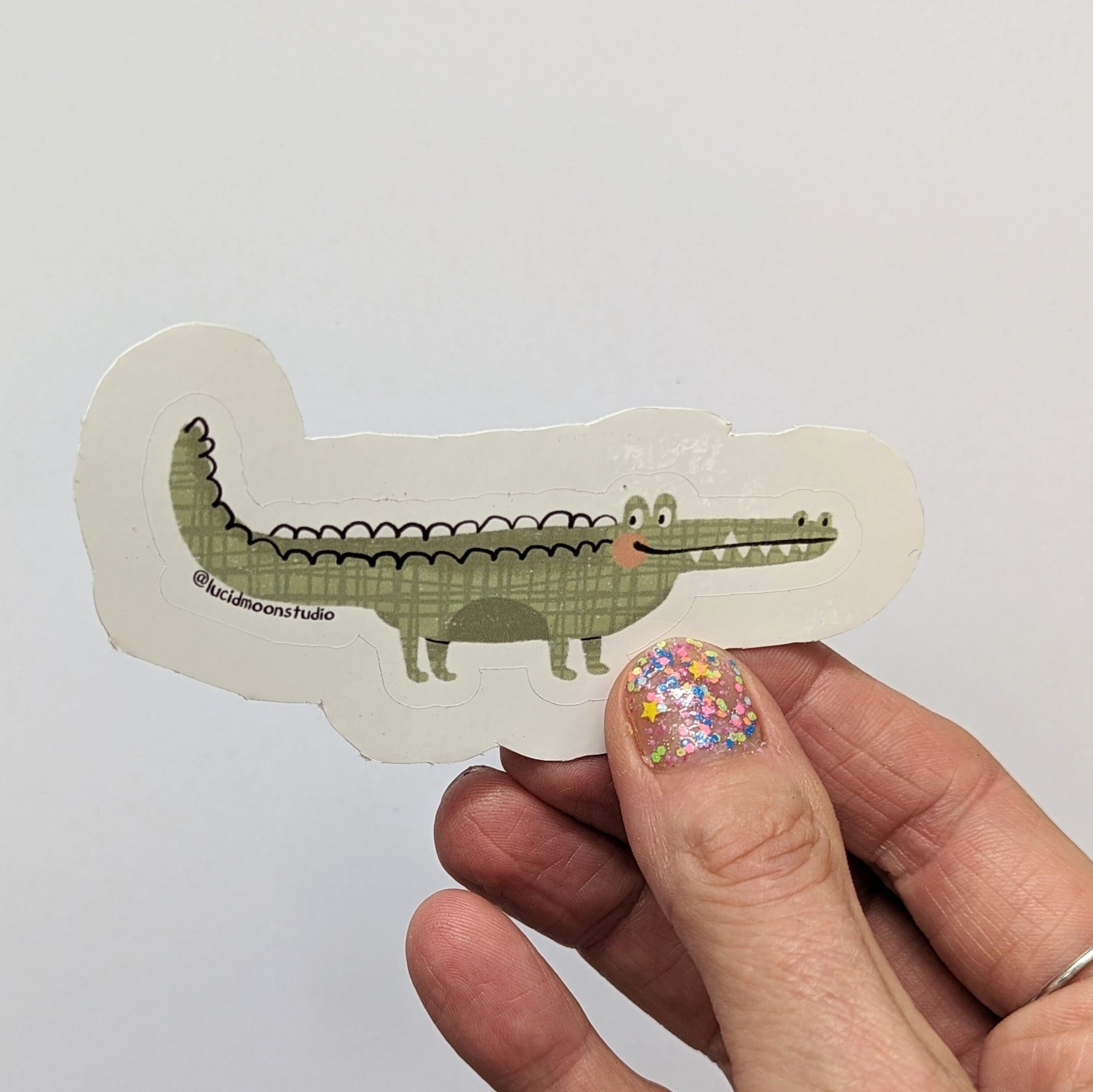 A hand holding a glossy vinyl die cut sticker with a hand drawn illustration of a cute green crocodile.
