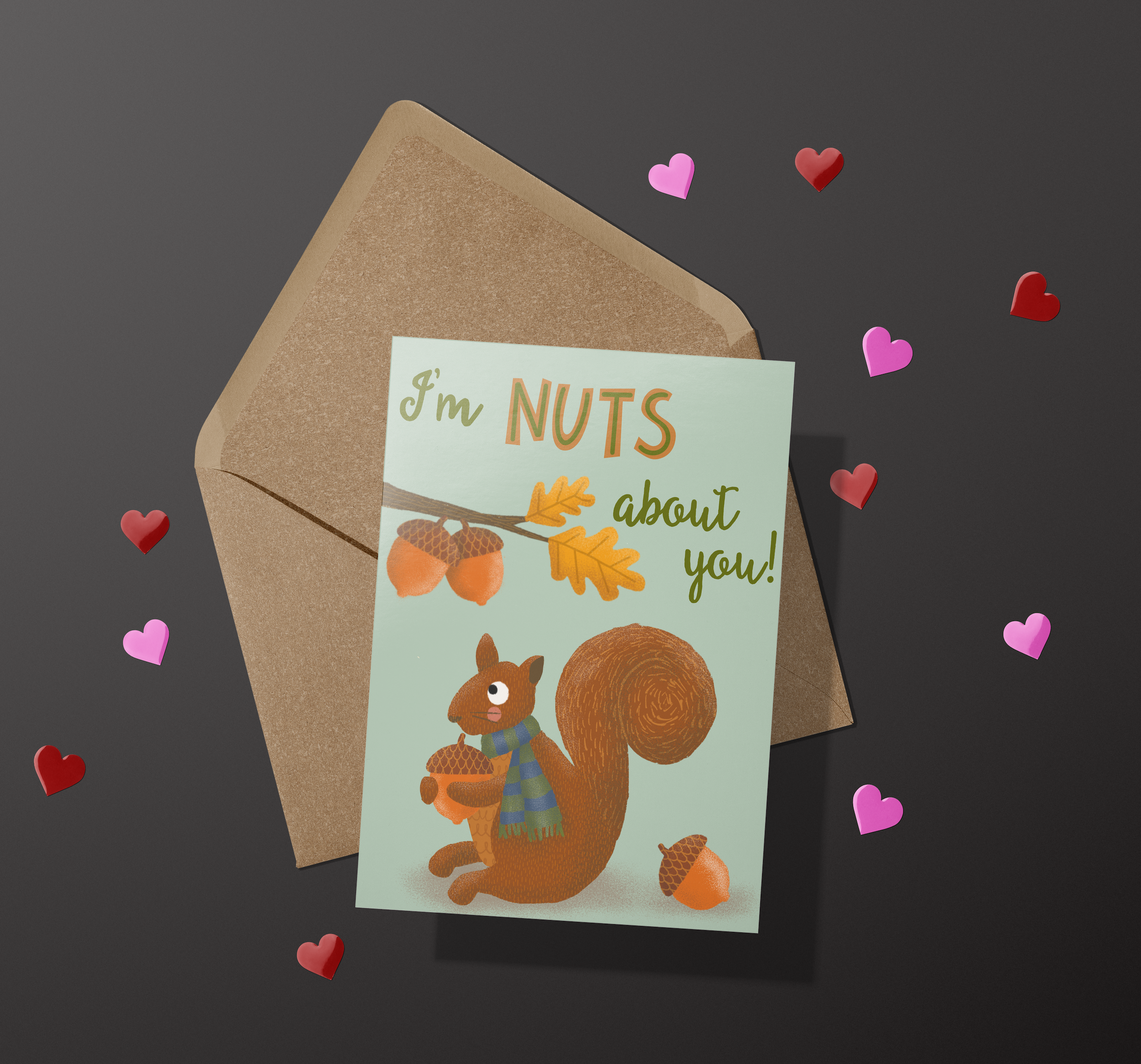 A greeting card with an illustration of a squirrel holding an acorn and the words I'm Nuts About You! on a kraft envelope on a black background with red and pink foil hearts