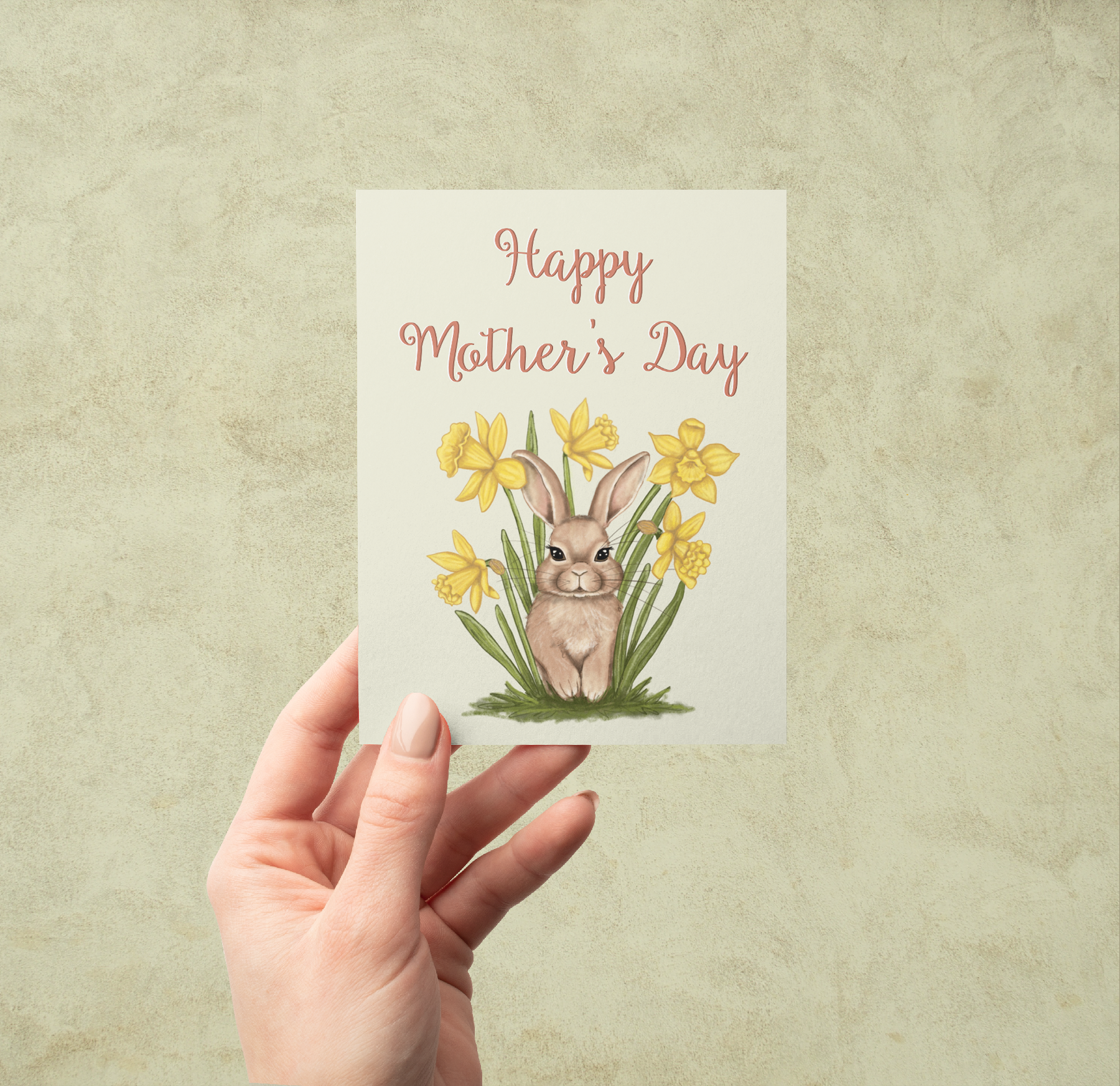 A hand holding a greeting card with a hand-painted bunny in daffodils and the words Happy Mother's Day against a muted background
