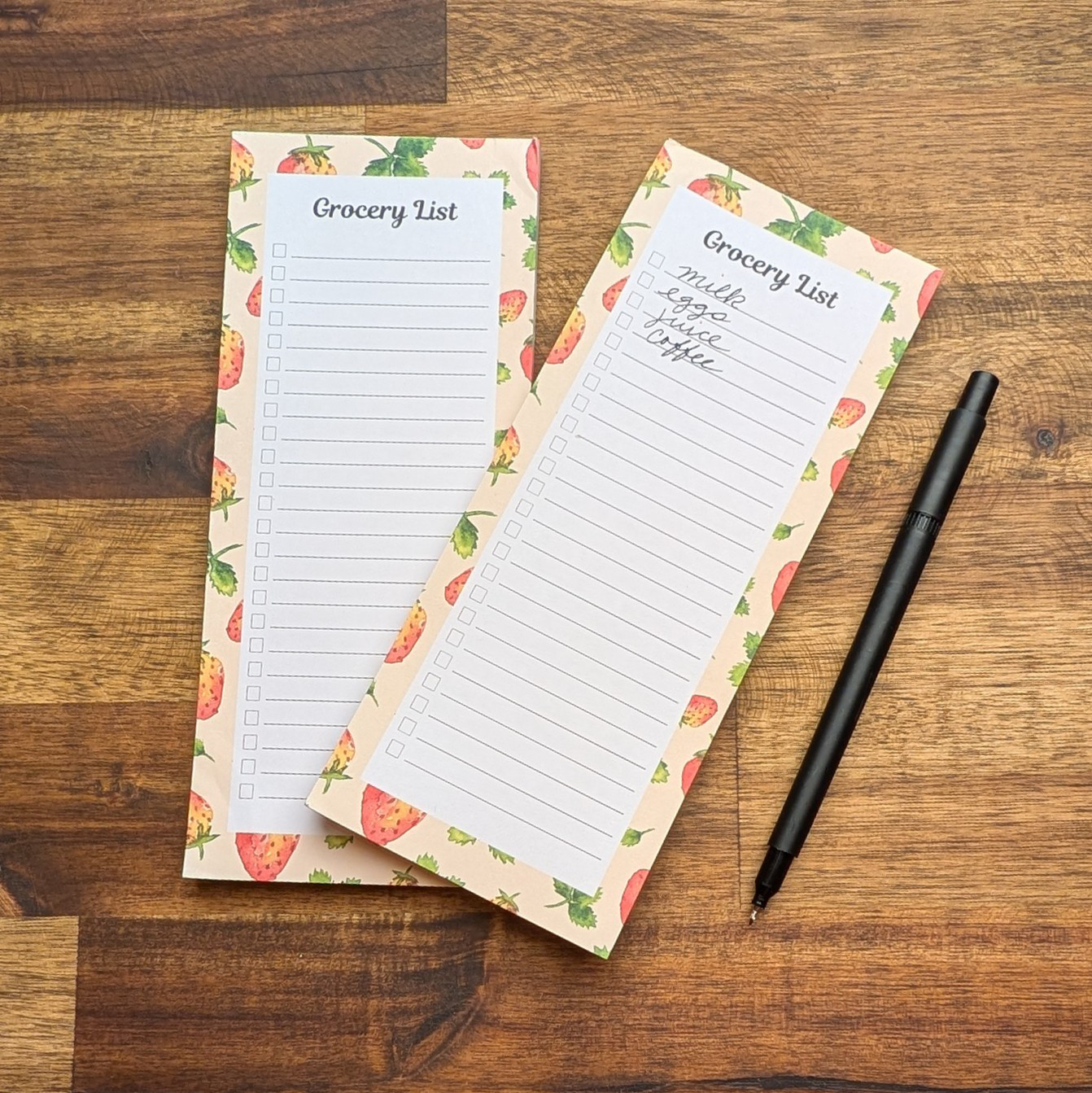 Two checklist grocery list notepads with a hand-painted watercolor strawberry border on a wood table with a black pen