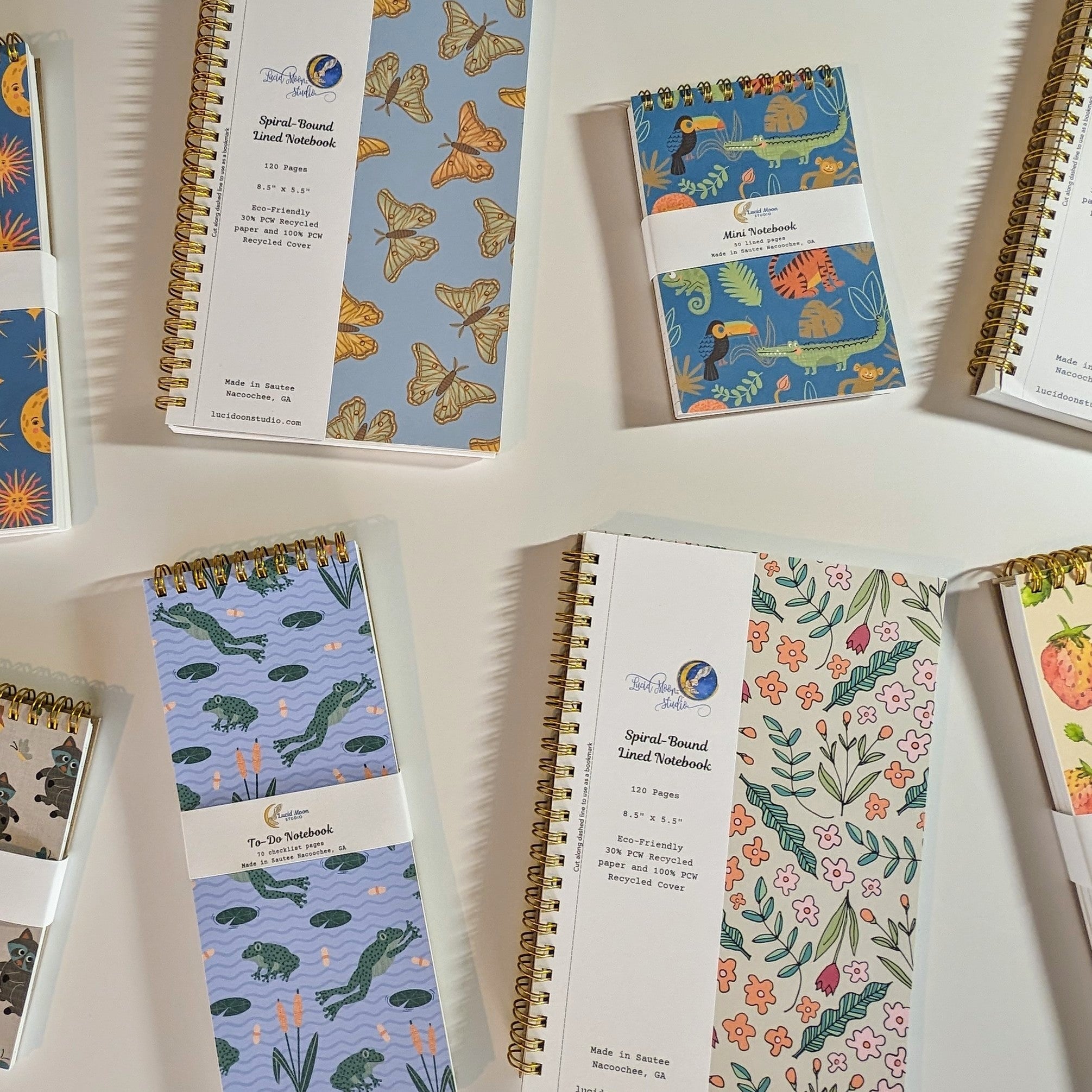 An assortment of Lucid Moon Studio spiral notebooks with colorful nature-inspired patterns on the covers on a white background