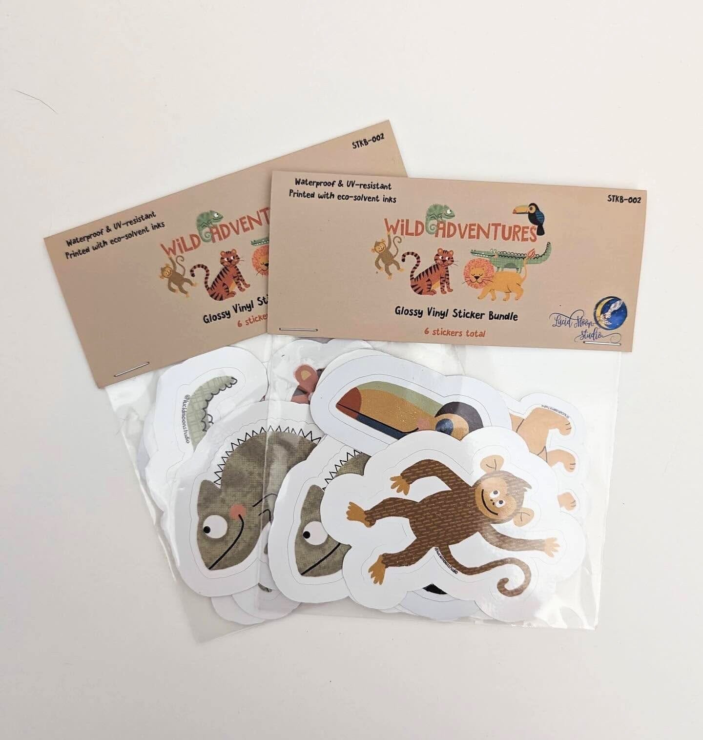 A photo of 2 Wild Adventures sticker bundle packs, each containing 6 glossy waterproof vinyl sticker designs that include a crocodile, chameleon, monkey, toucan, lion, and tiger packaged in a clear PLA certified compostable bag with cardstock label.