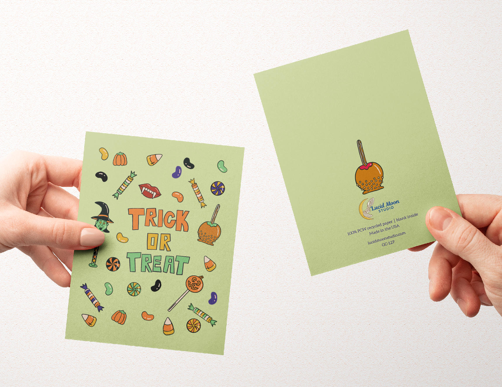 Two hands hold up a light green greeting card and its back side against a plain white background. The front of the card features colorful Halloween-themed illustrations and the words 'TRICK OR TREAT' in orange.