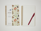 Gold spiral notebook with nature-themed cover design featuring mushrooms, butterflies, snails, frogs, blackberry branches, and bluebell flowers on a beige background and an open notebook showing white lined pages with a pen.