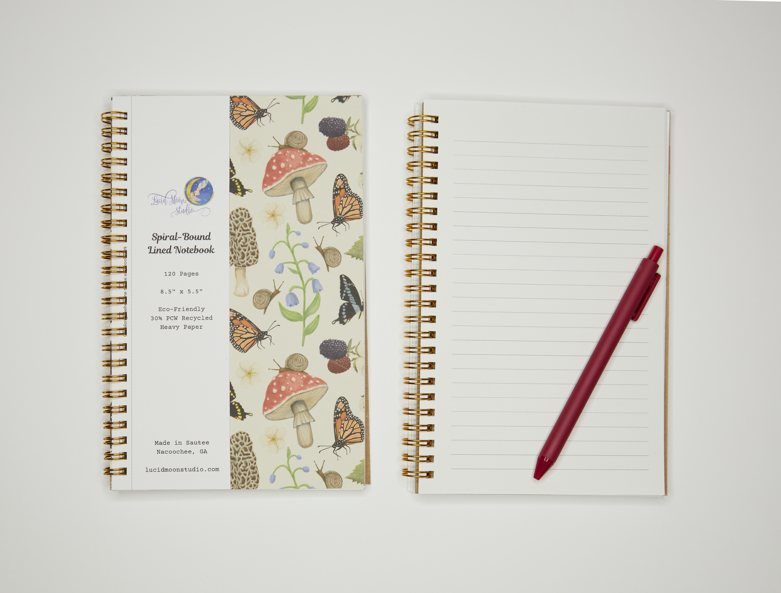 Gold spiral notebook with nature-themed cover design featuring mushrooms, butterflies, snails, frogs, blackberry branches, and bluebell flowers on a beige background and an open notebook showing white lined pages with a pen.