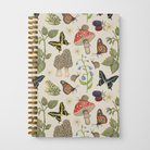 Gold spiral notebook with nature-themed cover design featuring mushrooms, butterflies, snails, frogs, blackberry branches, and bluebell flowers on a beige background.