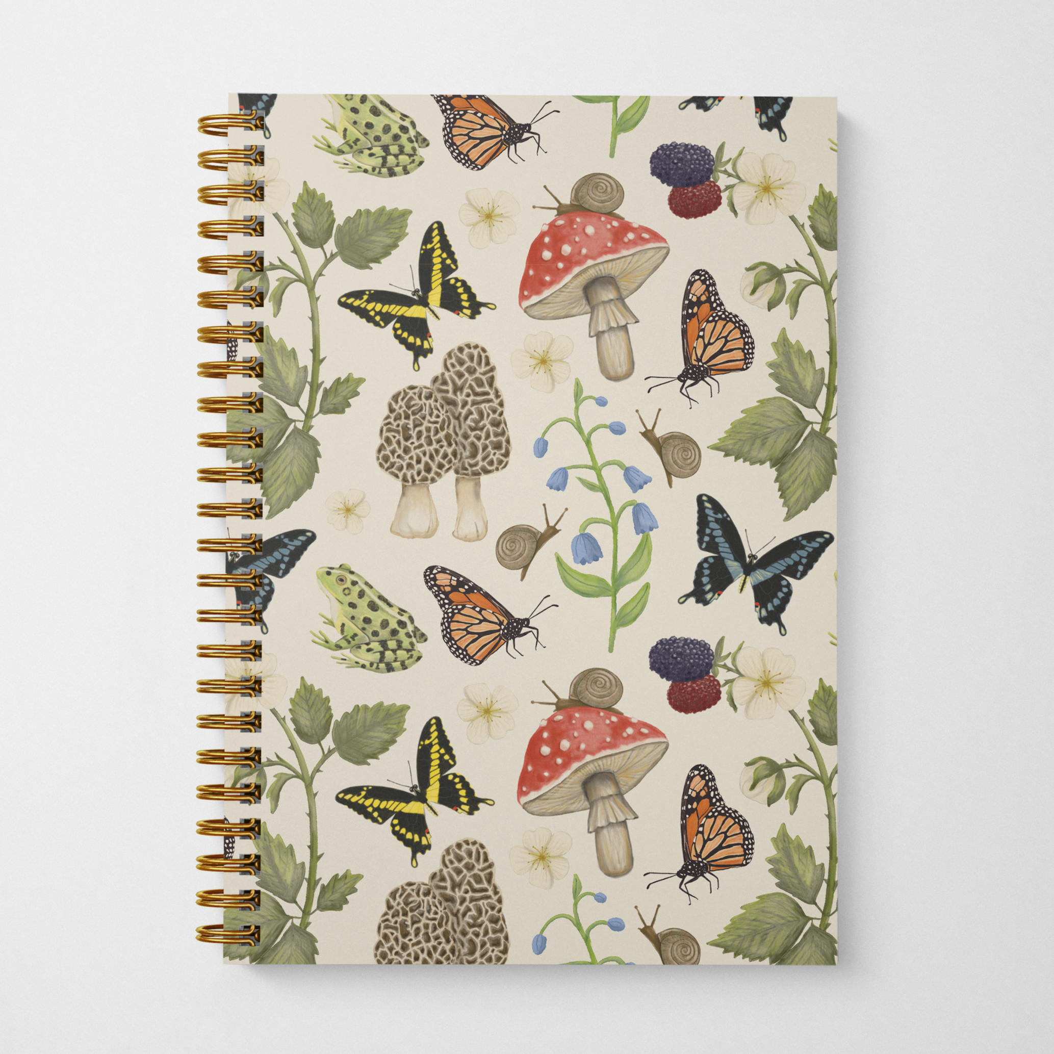 Gold spiral notebook with nature-themed cover design featuring mushrooms, butterflies, snails, frogs, blackberry branches, and bluebell flowers on a beige background.